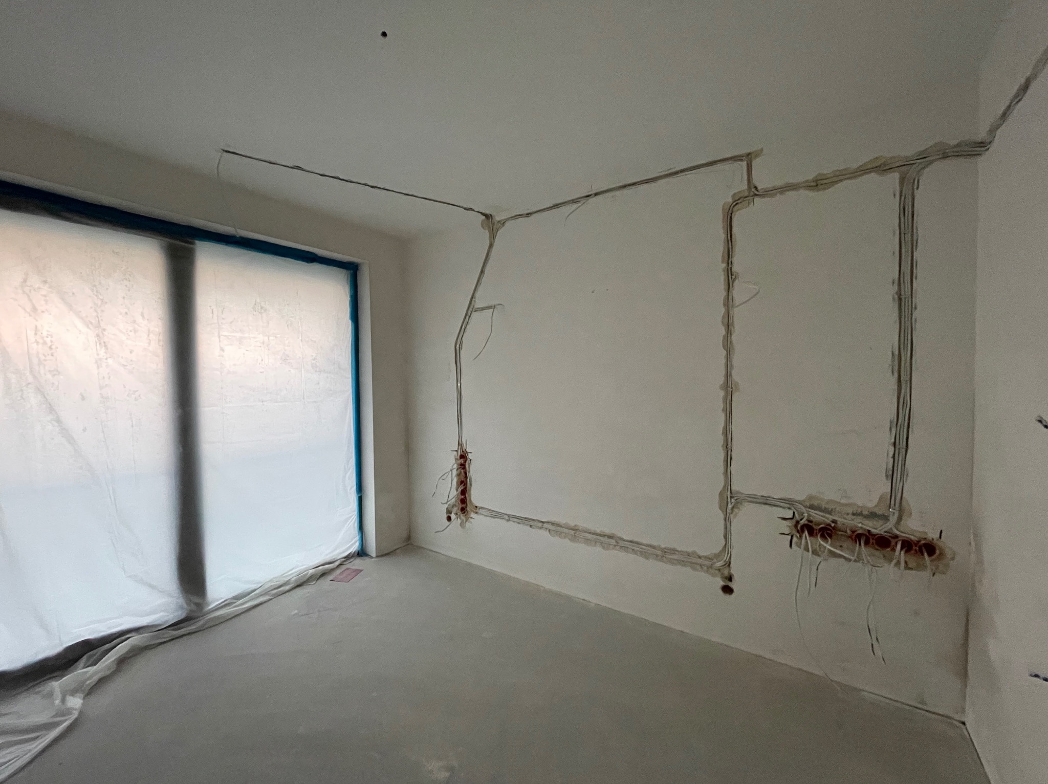 Wnętrze w trakcie remontu: widoczne bruzdy w ścianach z poprowadzonymi kablami elektrycznymi i puszkami instalacyjnymi, okno zabezpieczone folią malarską.