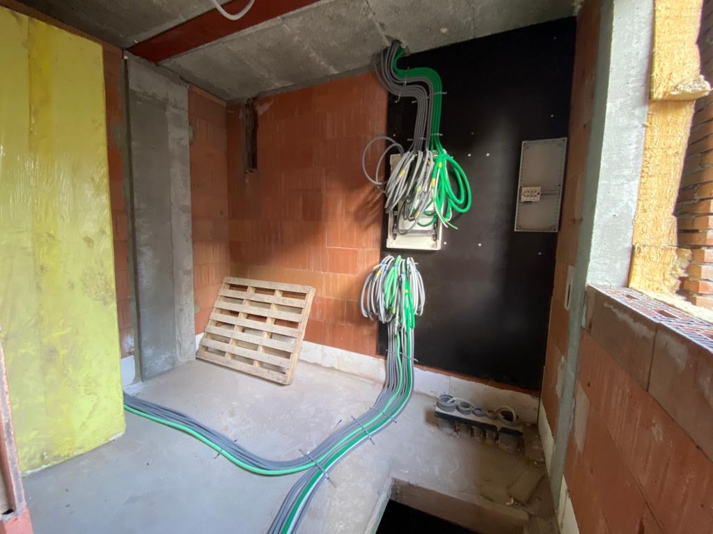 Surowy stan: Kable elektryczne w peszlach, częściowo podłączone do skrzynki na czarnej płycie w pomieszczeniu z ceglanymi ścianami i betonowym sufitem, widoczna paleta drewniana i żółta płyta...