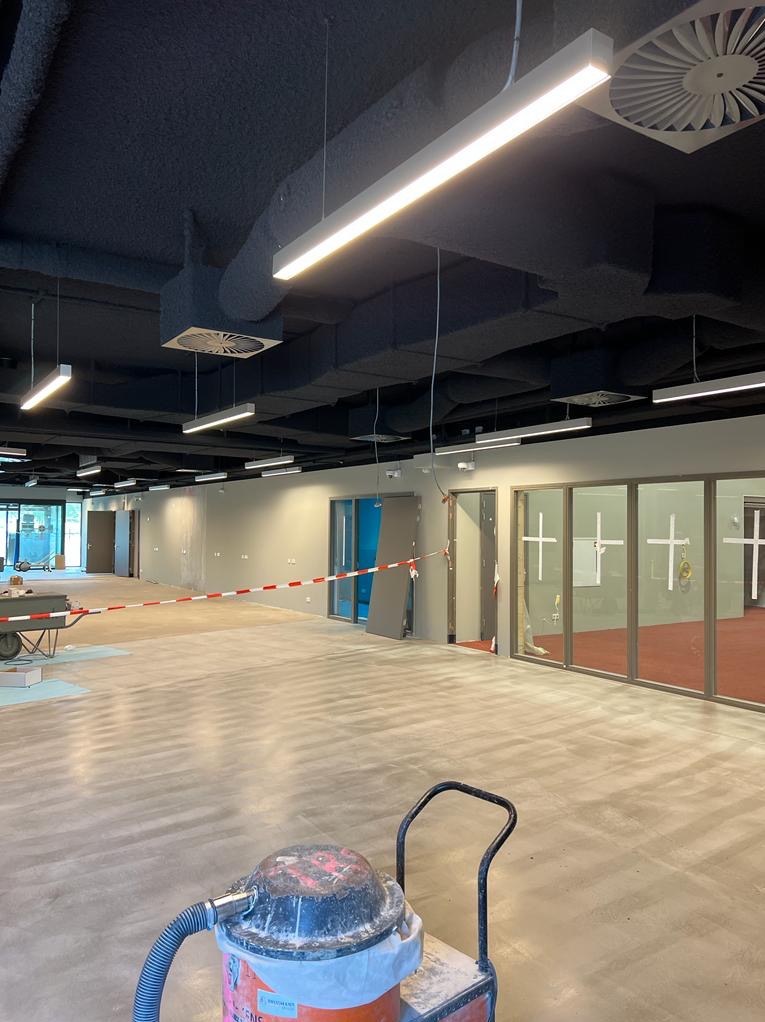 Wnętrze w trakcie wykańczania: widoczne lampy LED pod sufitem z instalacją wentylacyjną, taśma ostrzegawcza, oraz odkurzacz przemysłowy na pierwszym planie.