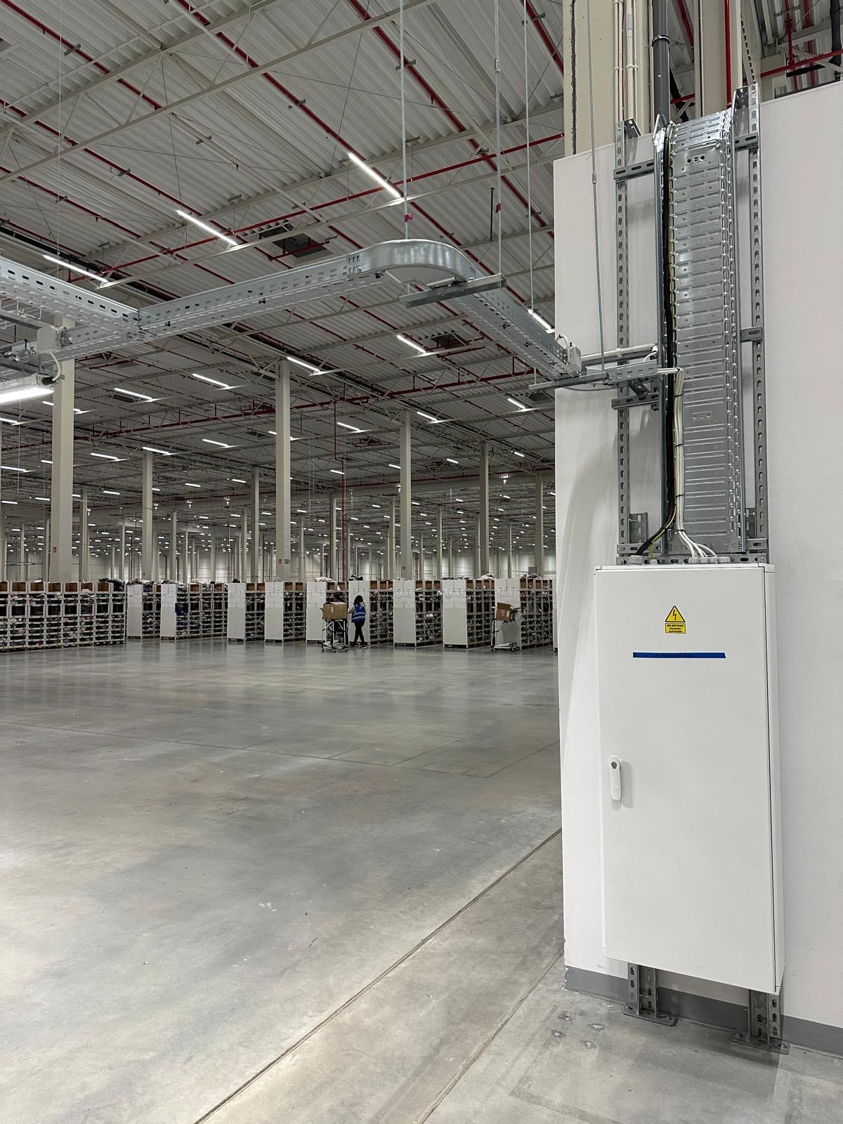 Rozległa hala magazynowa z zamontowanymi korytkami kablowymi pod sufitem i pionową trasą kablową na ścianie, zakończoną białą skrzynką elektryczną z symbolem ostrzegawczym, w tle wózki z kartonami...
