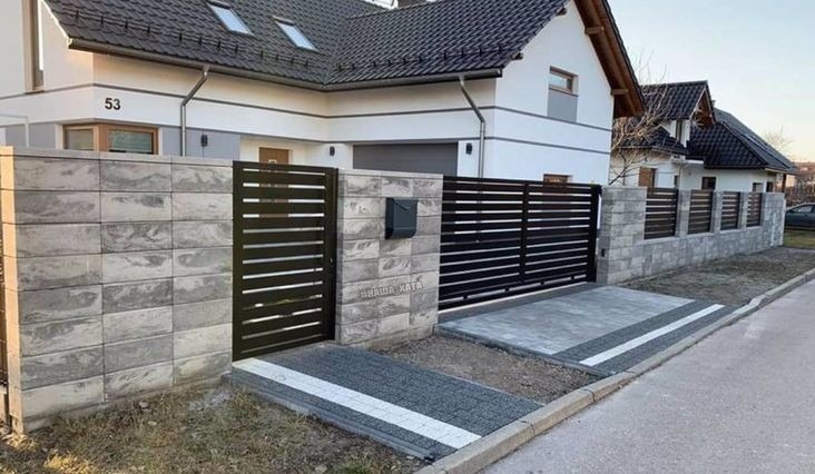 Nowoczesna posesja z bramą wjazdową i furtką z poziomych, czarnych profili, osadzonych w murze z szarej, imitującej kamień kostki, z widocznym numerem domu.