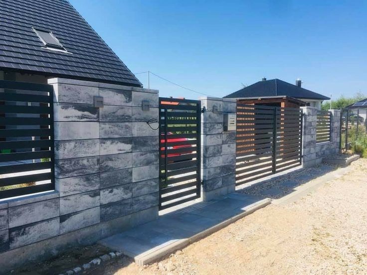 Nowoczesne ogrodzenie posesji z szarego betonu architektonicznego i poziomych, czarnych przęseł, z bramą wjazdową i furtką, na tle domu z ciemnym dachem i niebieskiego nieba.