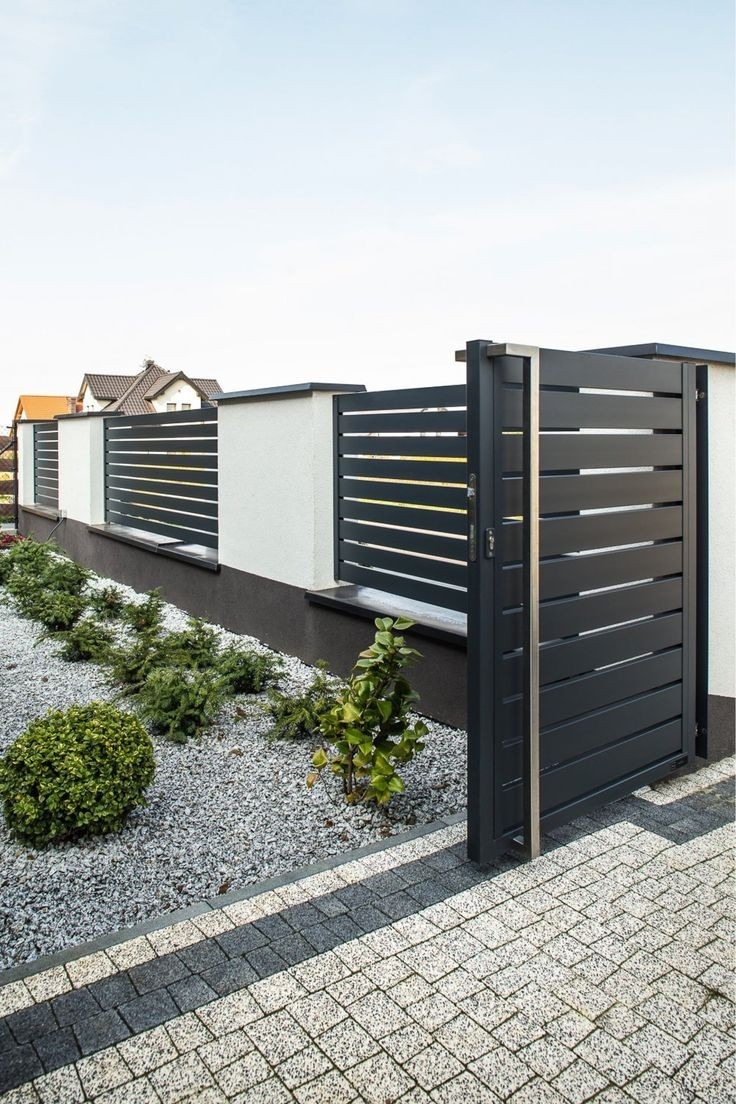 Nowoczesna, antracytowa brama wjazdowa z poziomymi panelami, częściowo otwarta, z widocznymi elementami stalowymi, osadzona w białym murku ogrodzeniowym. Ścieżka z kostki brukowej i ozdobny żwir...
