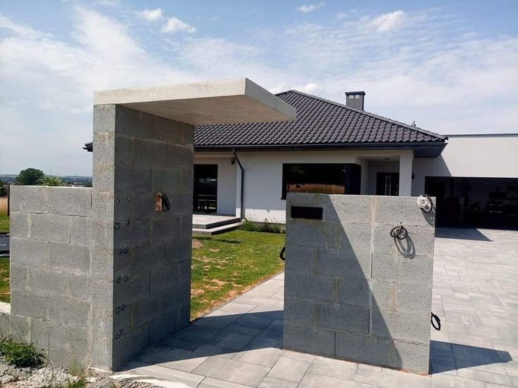 Surowa konstrukcja wejścia na posesję z szarego betonu komórkowego, z metalowymi uchwytami i betonowym daszkiem, na tle nowoczesnego domu z ciemną dachówką i podjazdem z kostki.