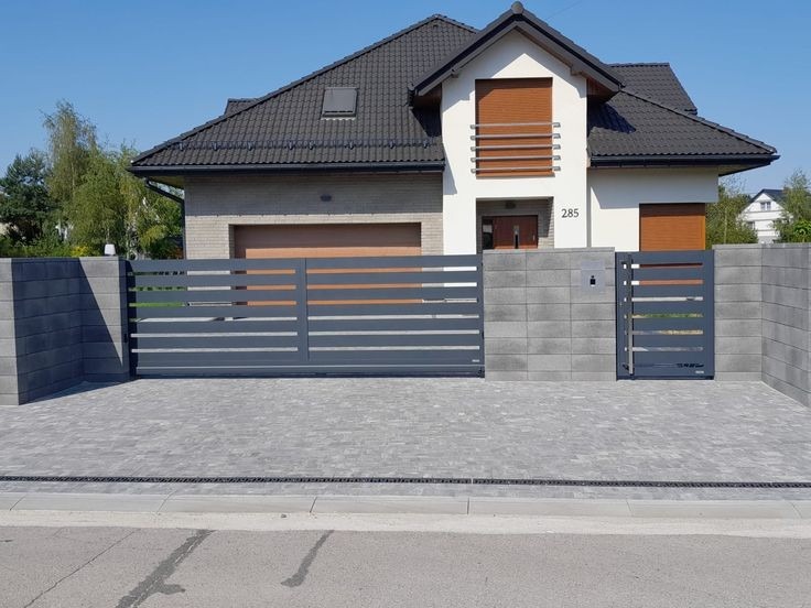 Nowoczesna posesja z przesuwną bramą wjazdową i furtką o poziomej konstrukcji, w kolorze grafitowym, na tle domu z jasną elewacją i szarą kostką brukową.