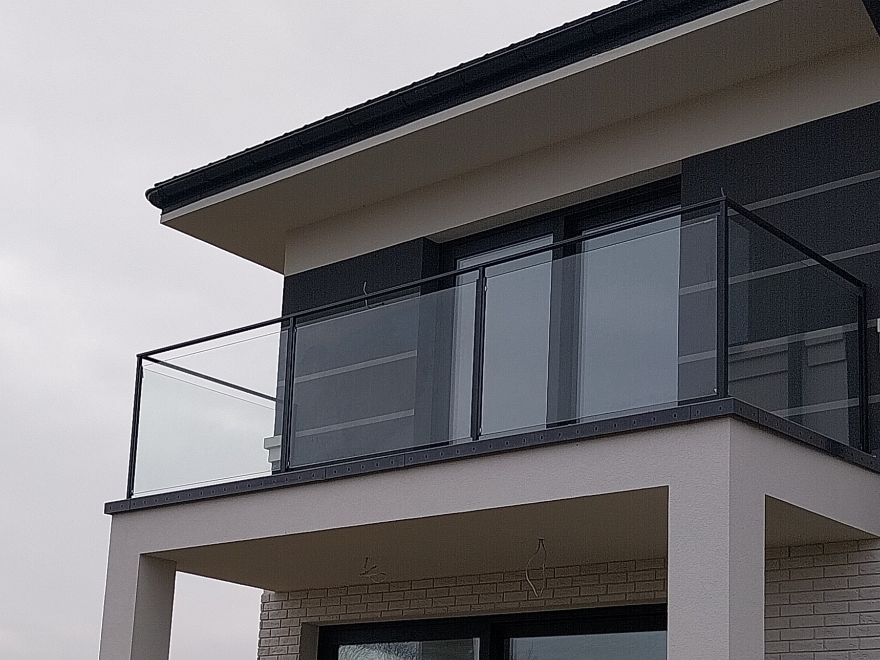 Nowoczesny balkon z szklaną balustradą i czarną ramą, widok z dołu na elewację budynku z elementami cegły.