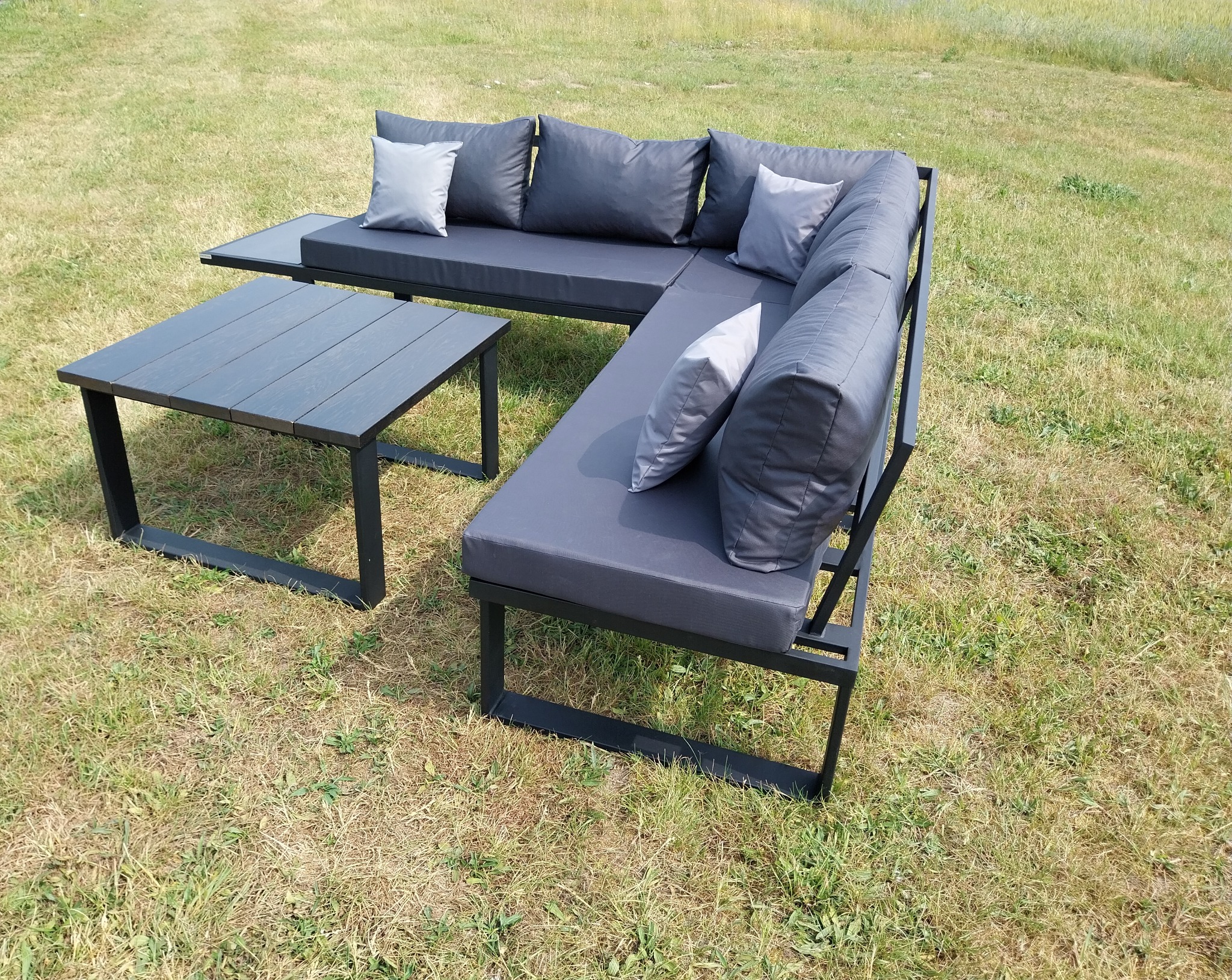Zestaw mebli ogrodowych: narożna sofa z szarego metalu z poduchami i stolik kawowy na trawniku.