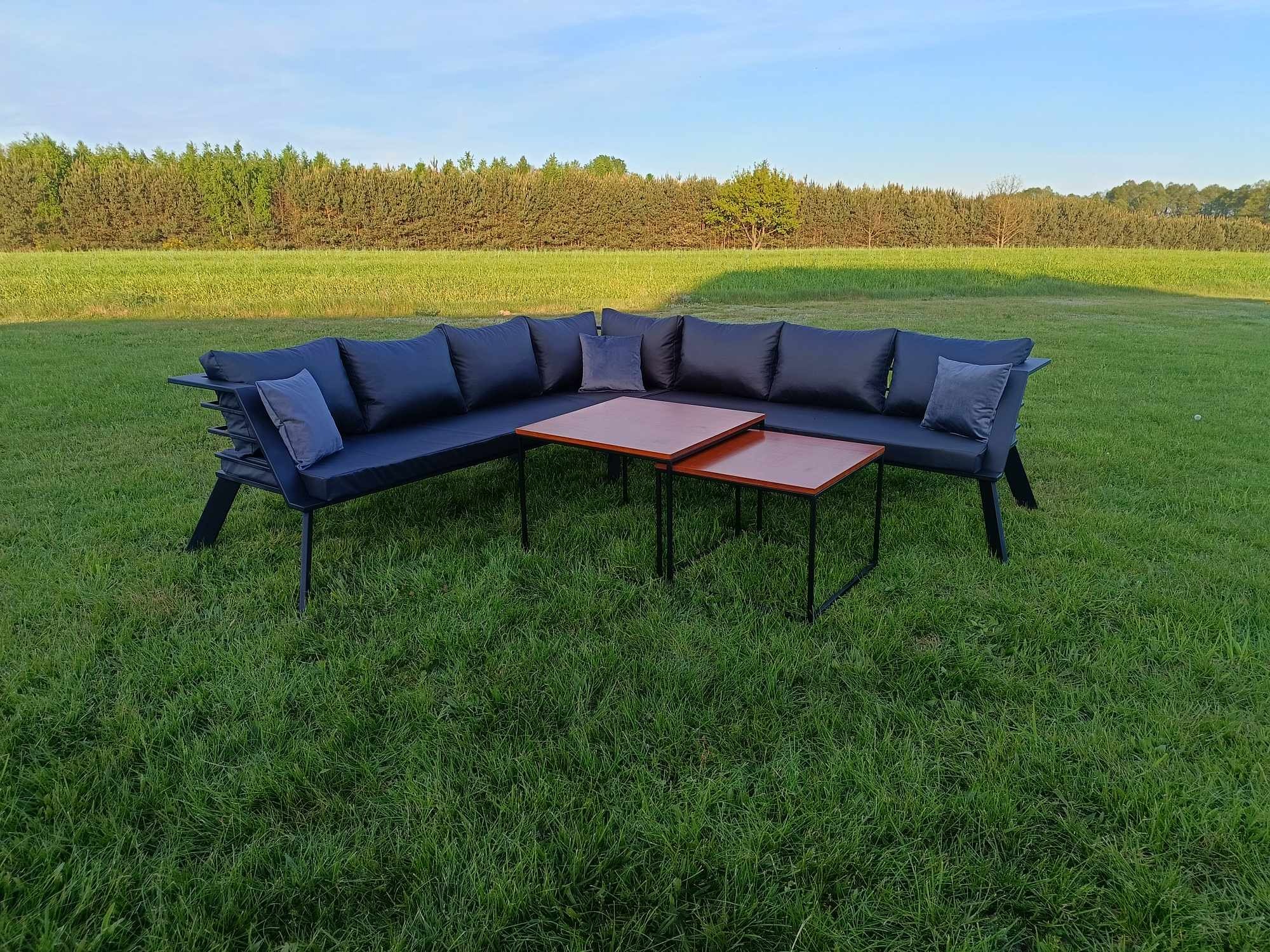 Duży narożnik z ciemnymi poduchami i dwa stoliki kawowe na trawniku przed lasem.