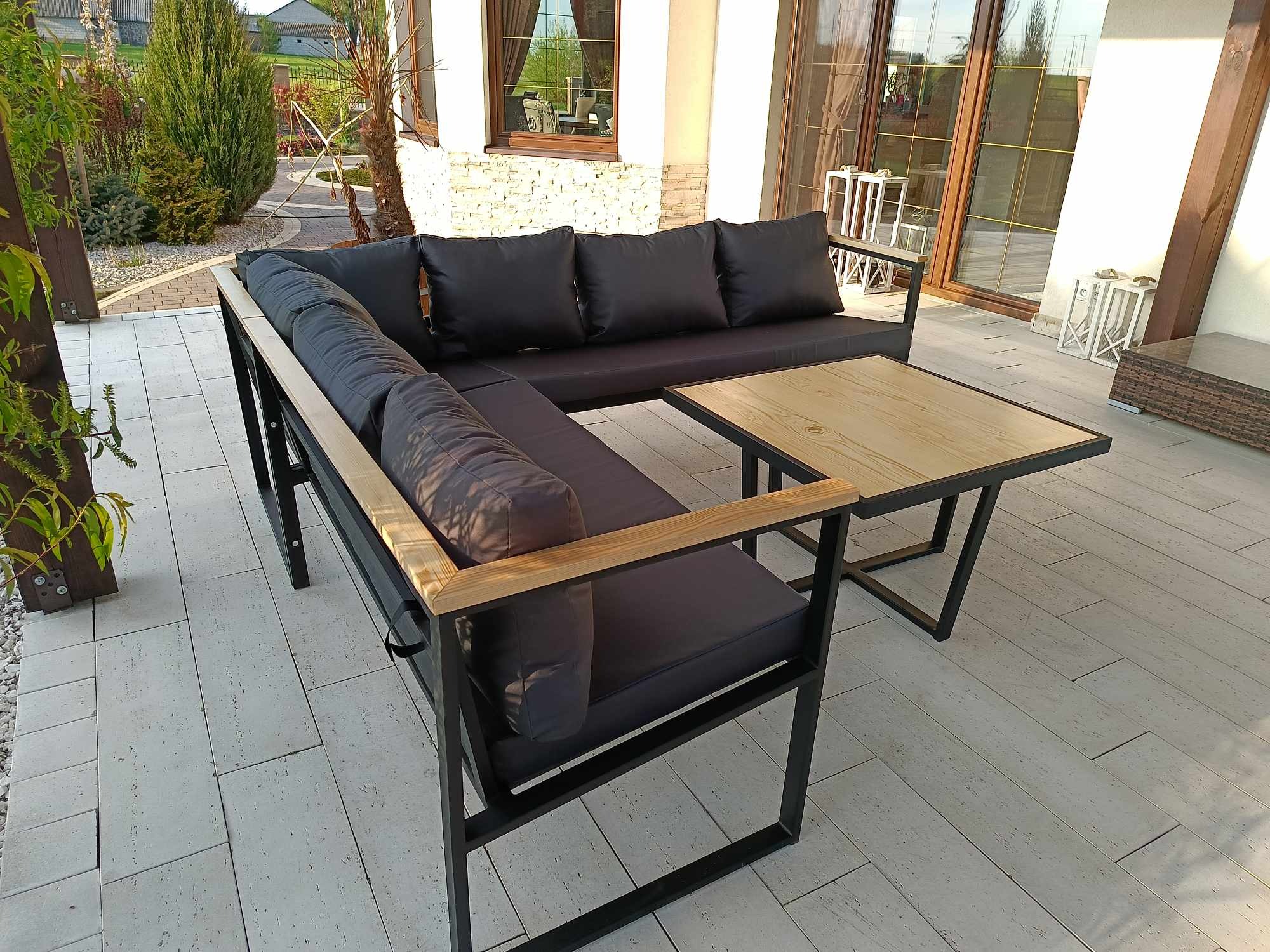 Zestaw mebli ogrodowych: narożna sofa z czarnymi poduszkami i drewnianymi podłokietnikami oraz stolik kawowy z drewnianym blatem i czarną metalową ramą, na tarasie z jasnych płyt.