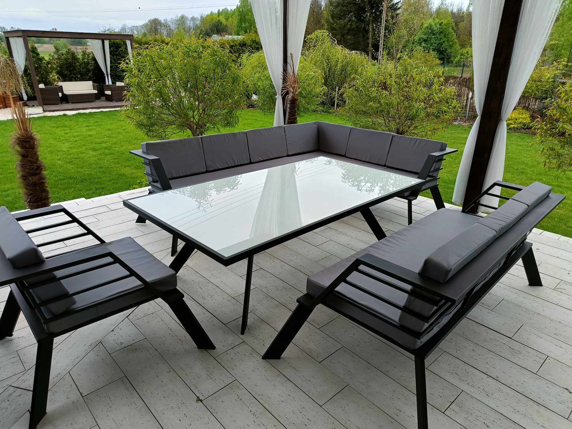 Zestaw mebli ogrodowych z szarego metalu i jasnymi poduchami, ustawiony na tarasie wyłożonym płytkami imitującymi drewno, w tle trawnik i altana z zasłonami.