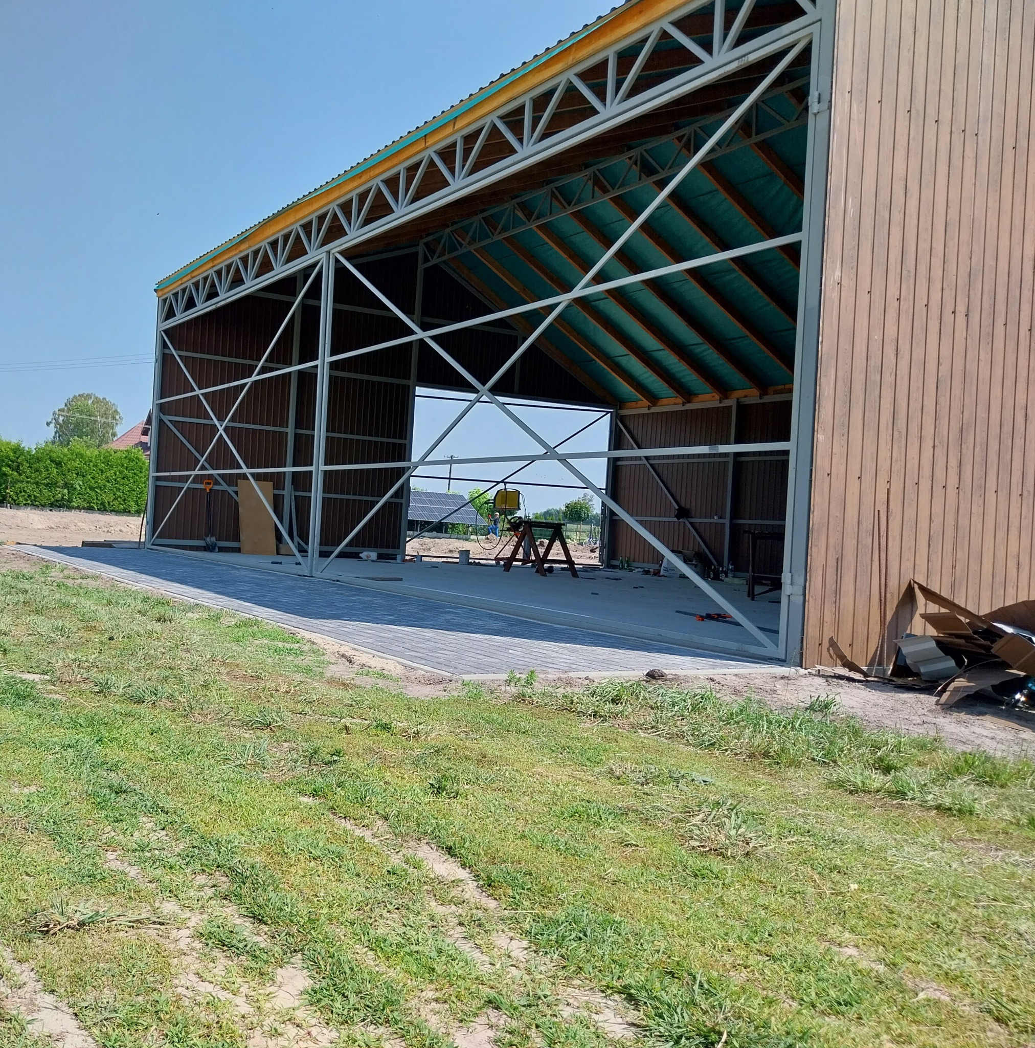 Konstrukcja stalowa hali w trakcie budowy, widoczny szkielet i częściowo obłożone ściany z desek, na podłodze rozłożone narzędzia i materiały budowlane, w tle dom z panelami słonecznymi.