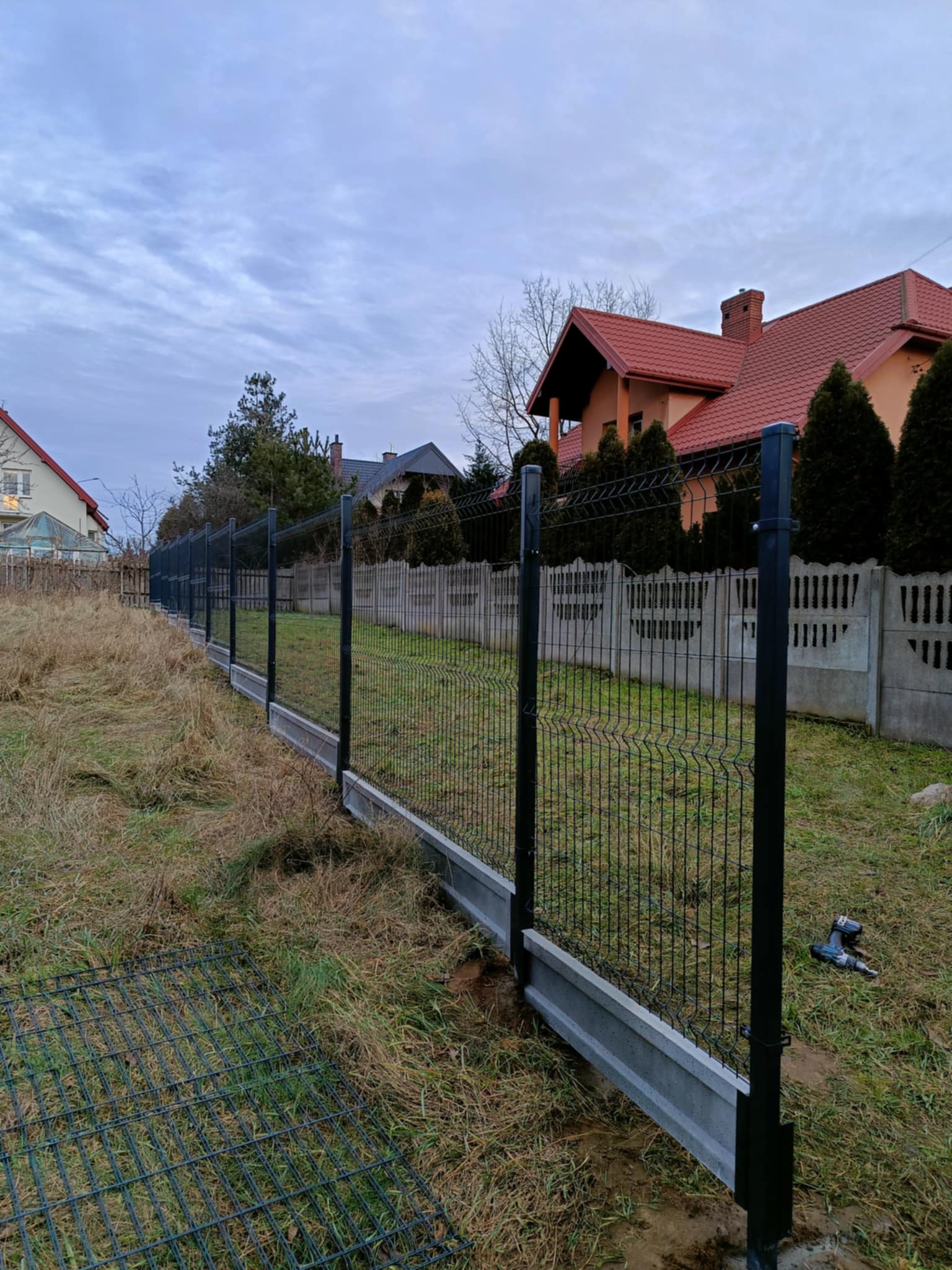 Częściowo zamontowane ogrodzenie panelowe z podmurówką betonową na tle domów i pochmurnego nieba. W trawie leży fragment siatki i widać wkrętarkę.
