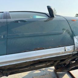 Maciej Grzęda MG Serwis - Zielony samochód na podnośniku, widoczne ślady korozji na progach i naprawiane blacharskie elementy karoserii, z naklejką z kodem kreskowym.