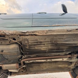 Maciej Grzęda MG Serwis - Silnie skorodowane podwozie samochodu osobowego z widocznymi elementami układu wydechowego, zbiornika paliwa i zawieszenia, sfotografowane pod kątem od dołu.