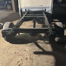 Maciej Grzęda MG Serwis - Rama samochodu dostawczego po konserwacji antykorozyjnej, widok z tyłu, czarna, na betonowej posadzce warsztatu, z widocznymi elementami karoserii i kołami.