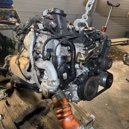 Maciej Grzęda MG Serwis - Wyjęty silnik samochodowy zawieszony na łańcuchu w warsztacie, widoczne przewody, rury i osprzęt, obok leży wkrętarka.