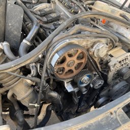 Maciej Grzęda MG Serwis - Otwarty silnik samochodowy z widocznym rozrządem, paskiem zębatym i kołami pasowymi w zbliżeniu, elementy lekko zabrudzone.