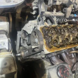 Otwarty silnik samochodowy z widocznym paskiem rozrządu, wałkami rozrządu i innymi elementami wewnętrznymi, pokrytymi olejem. Widoczny brud i zużycie na komponentach.