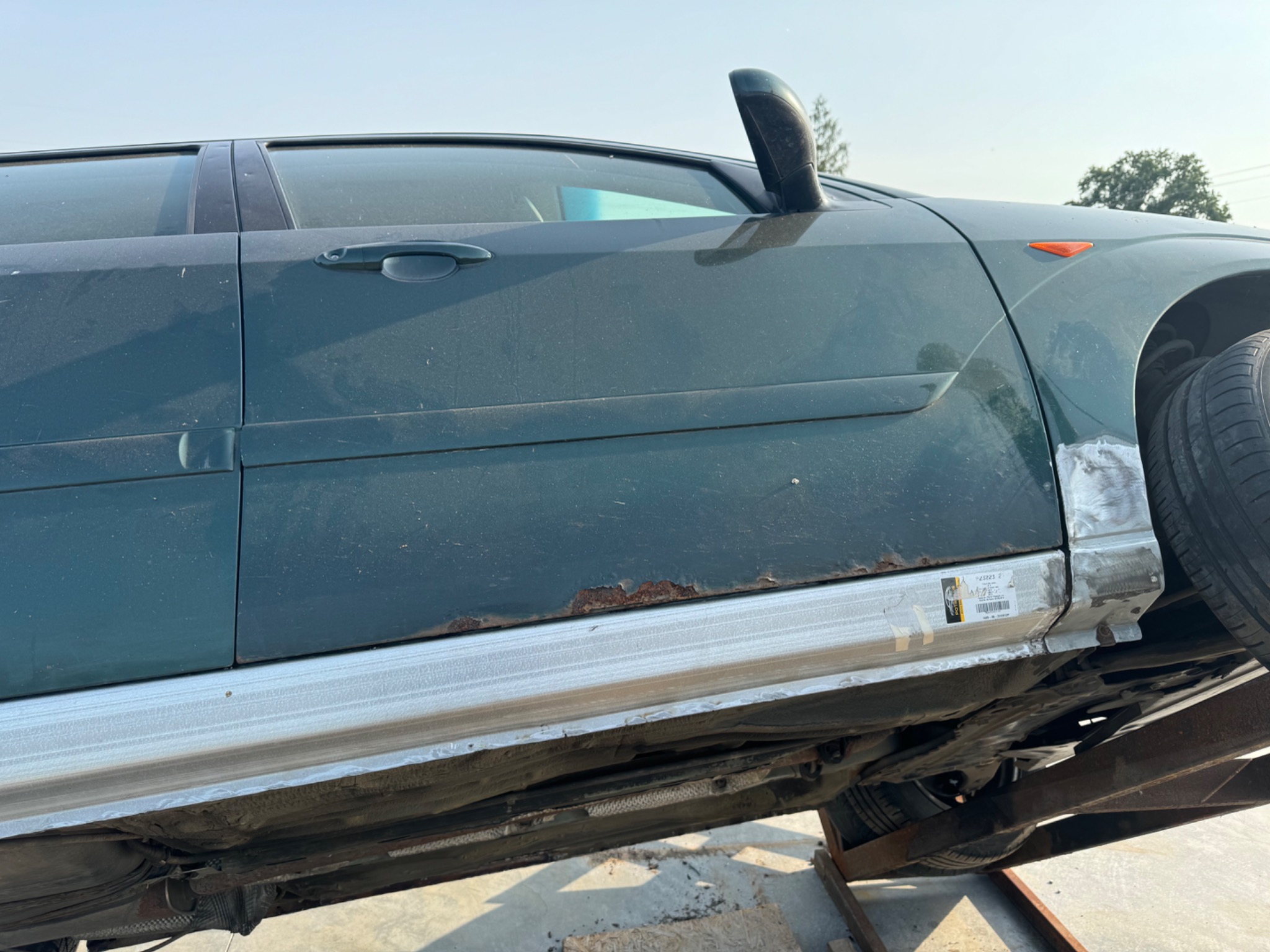 Zielony samochód na podnośniku, widoczne ślady korozji na progach i naprawiane blacharskie elementy karoserii, z naklejką z kodem kreskowym.