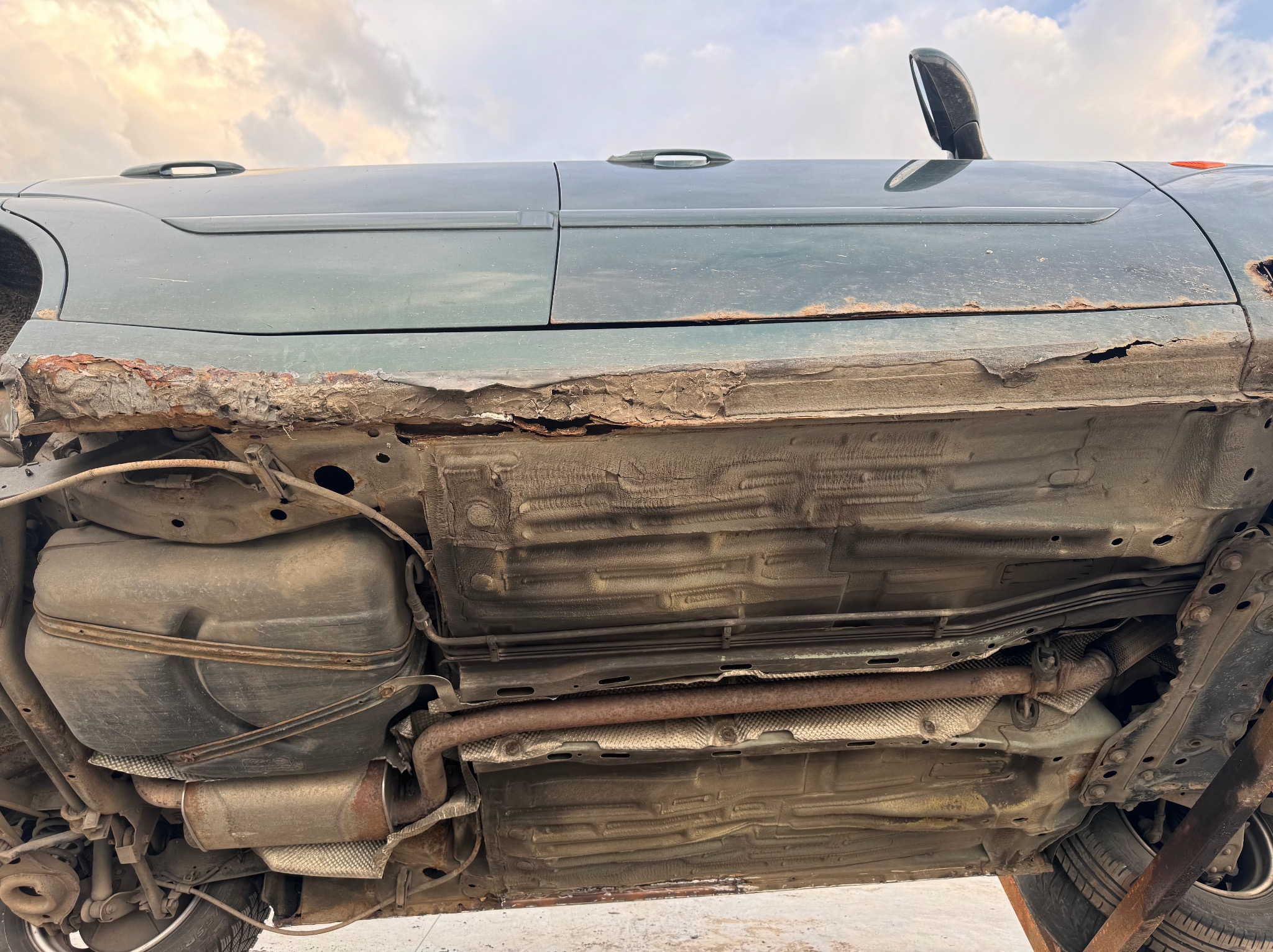 Silnie skorodowane podwozie samochodu osobowego z widocznymi elementami układu wydechowego, zbiornika paliwa i zawieszenia, sfotografowane pod kątem od dołu.