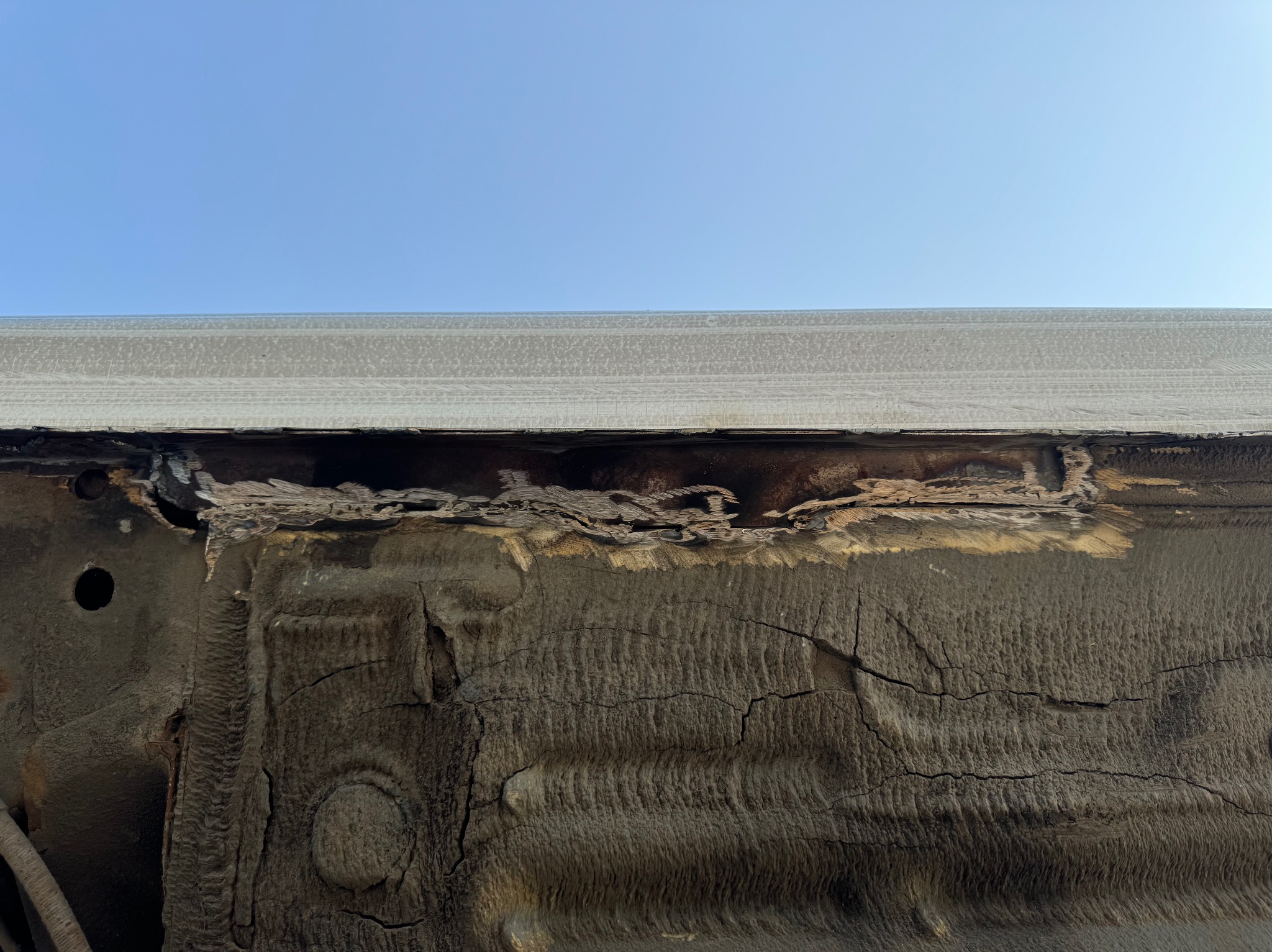 Silnie skorodowany fragment podwozia samochodu, widoczne liczne ogniska rdzy i uszkodzenia metalu, na tle błękitnego nieba.