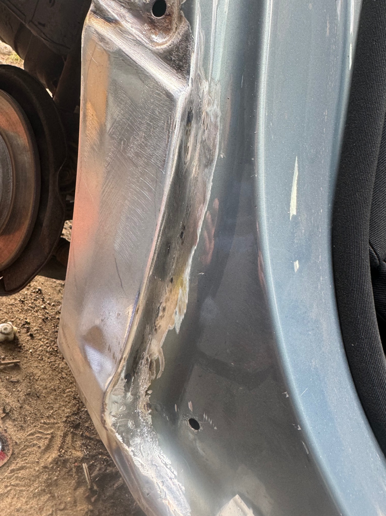 Fragment karoserii samochodu w trakcie naprawy blacharskiej, widoczne ślady szlifowania i spawania, przygotowanie do lakierowania.