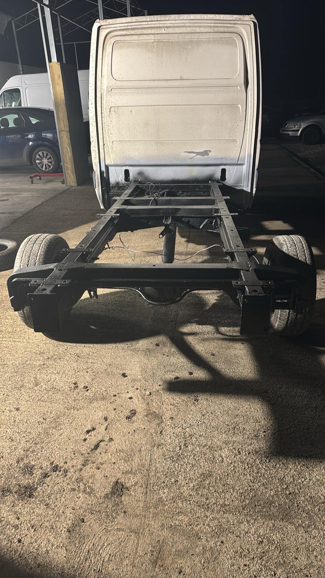 Rama samochodu dostawczego po konserwacji antykorozyjnej, widok z tyłu, czarna, na betonowej posadzce warsztatu, z widocznymi elementami karoserii i kołami.