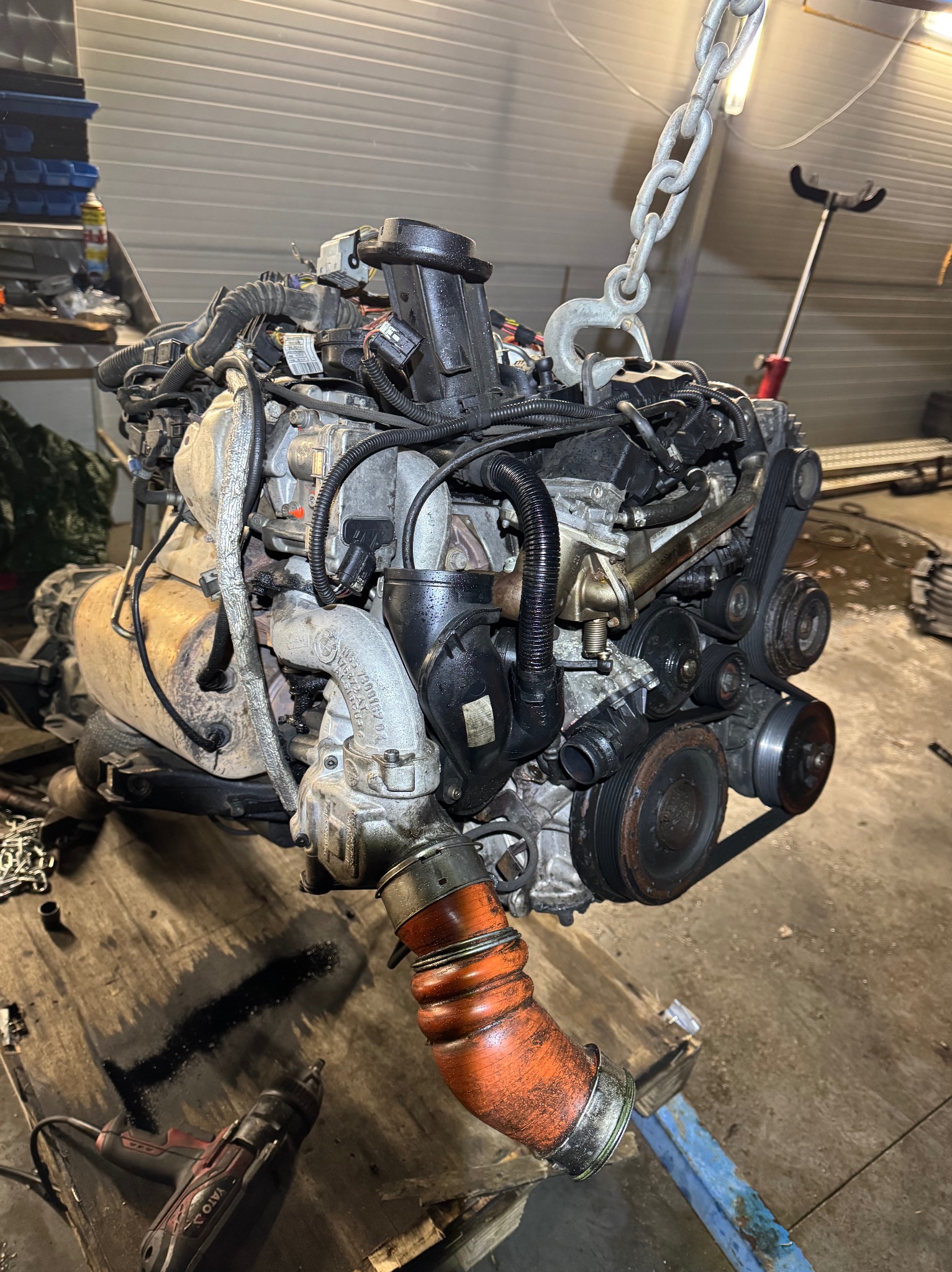Wyjęty silnik samochodowy zawieszony na łańcuchu w warsztacie, widoczne przewody, rury i osprzęt, obok leży wkrętarka.