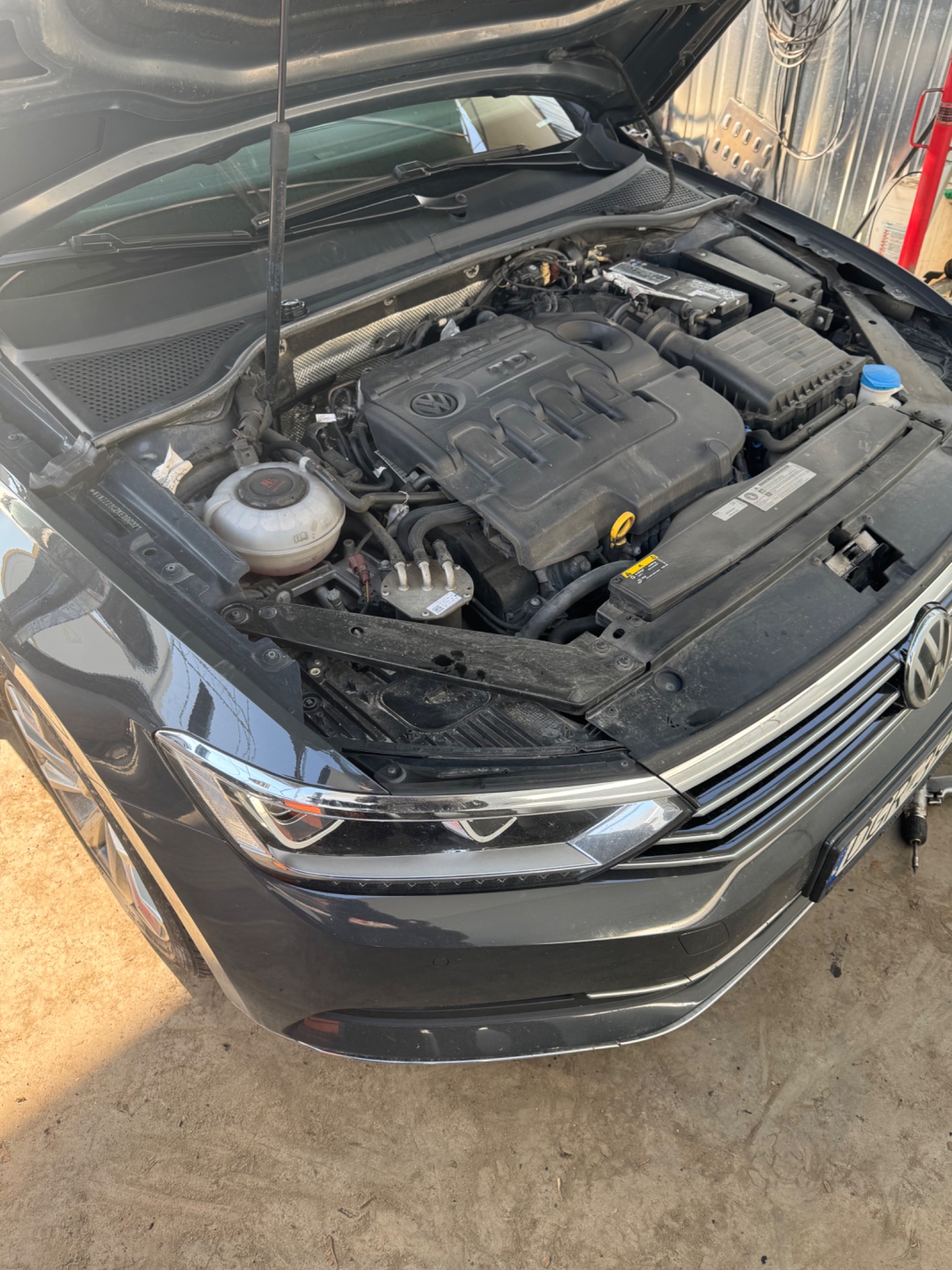 Otwarta maska szarego samochodu marki Volkswagen, widoczny silnik i elementy pod maską, warsztatowe tło.