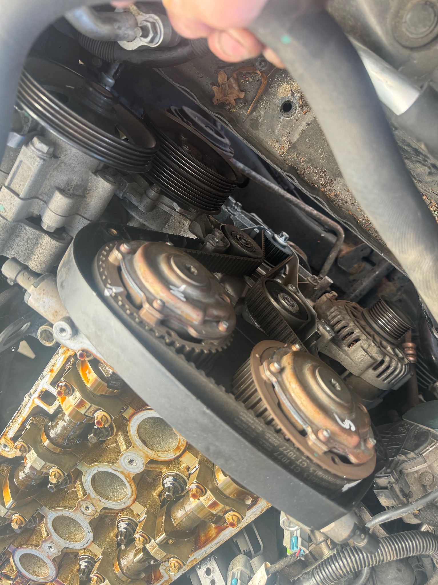 Widok z góry na otwarty silnik samochodowy podczas wymiany paska rozrządu, widoczne koła zębate, pasek, głowica cylindrów z olejem, elementy mocujące i fragment węża.