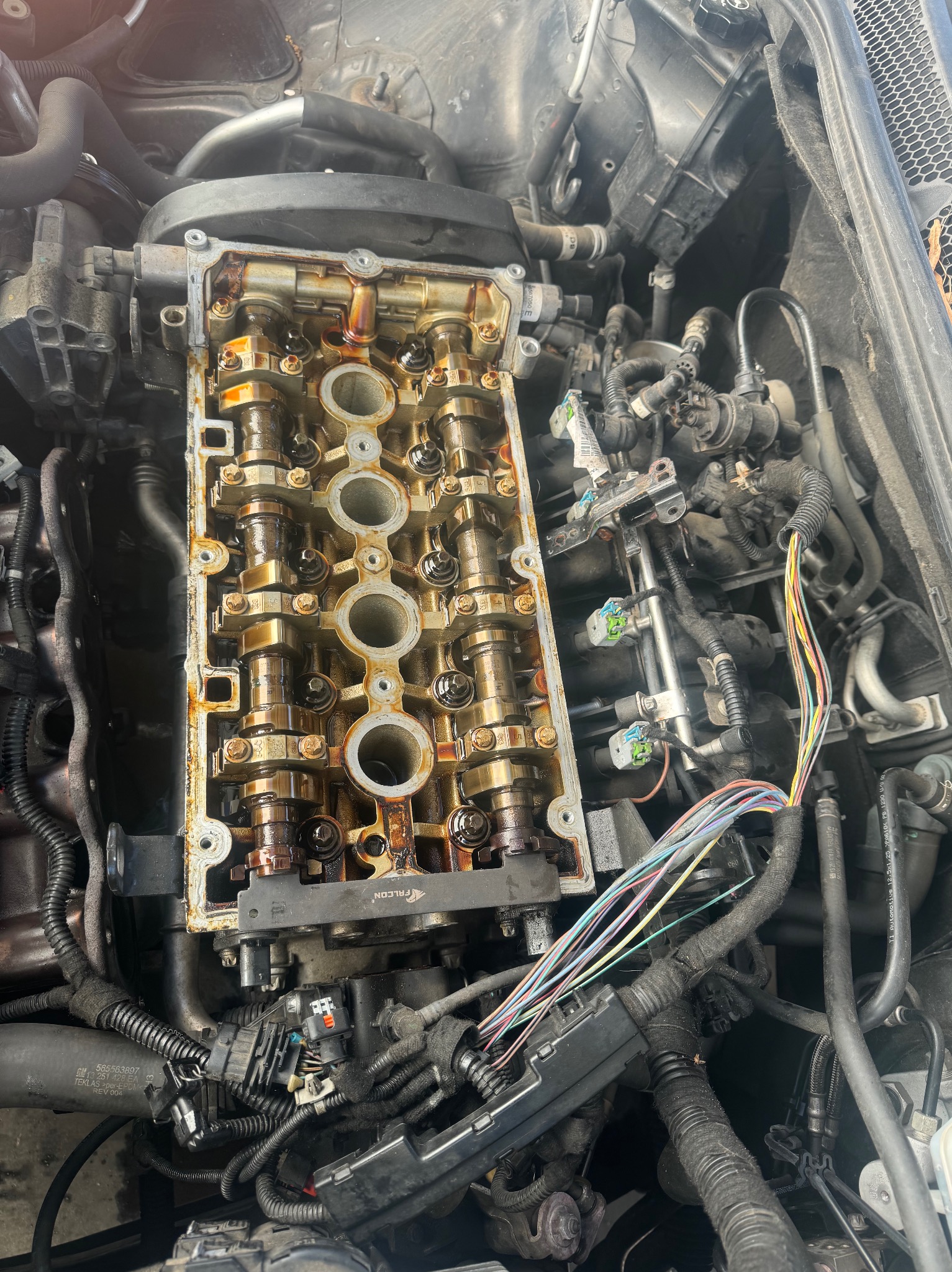 Otwarty silnik samochodowy z widoczną głowicą cylindrów, wałkami rozrządu i przewodami. Widoczne ślady oleju na elementach. Marka FALCON na elemencie. Skupienie na wnętrzu silnika.