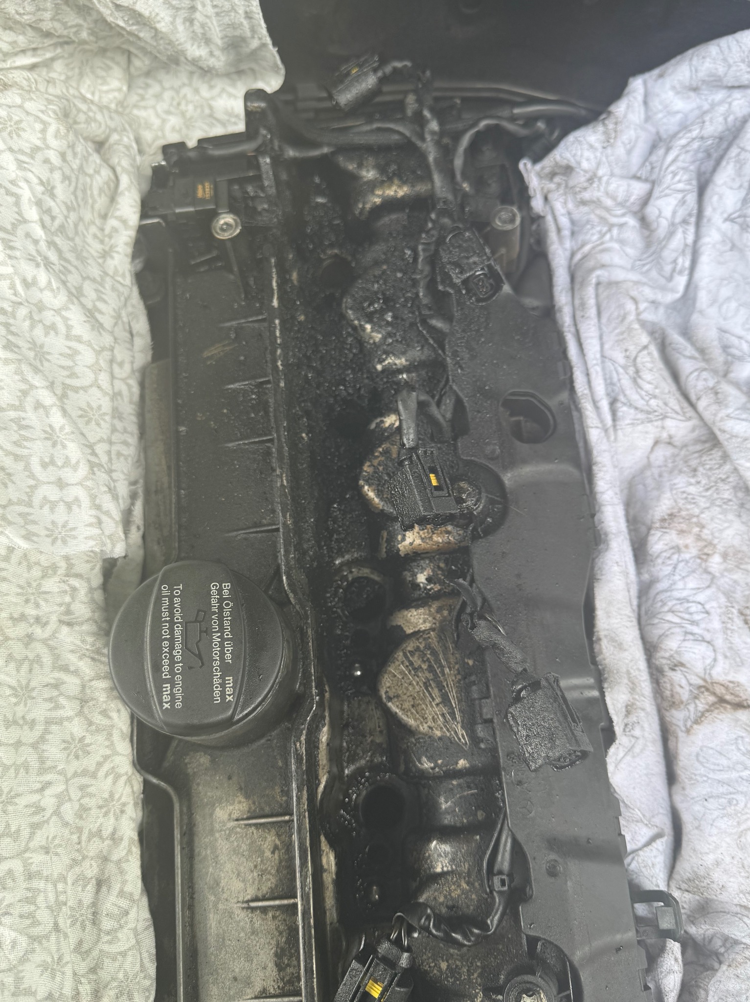 Zabrudzona olejem pokrywa zaworów silnika samochodowego, częściowo przykryta białą szmatką. Widoczne złącza elektryczne i napisy ostrzegawcze na korku wlewu oleju.