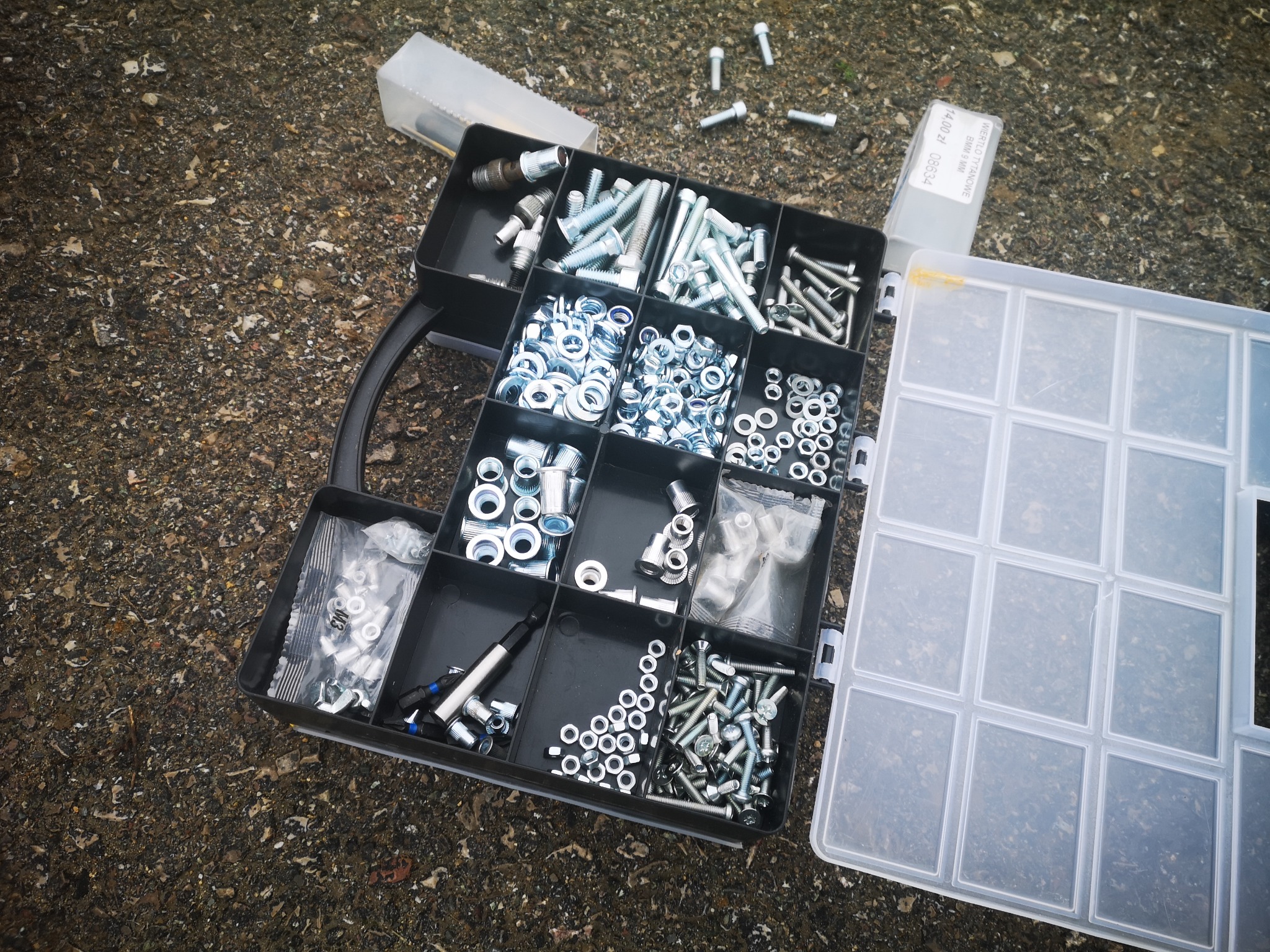 Otwarty organizer z przegródkami wypełnionymi różnymi śrubami, nakrętkami i podkładkami, leżący na szorstkiej, ciemnej powierzchni. Obok widoczny otwarty pojemnik z przezroczystego plastiku. W tle...