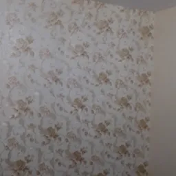 Ściana po tapetowaniu z beżowymi kwiatami, widoczne puszki z klejem i narzędzia malarskie na czarnej folii ochronnej.