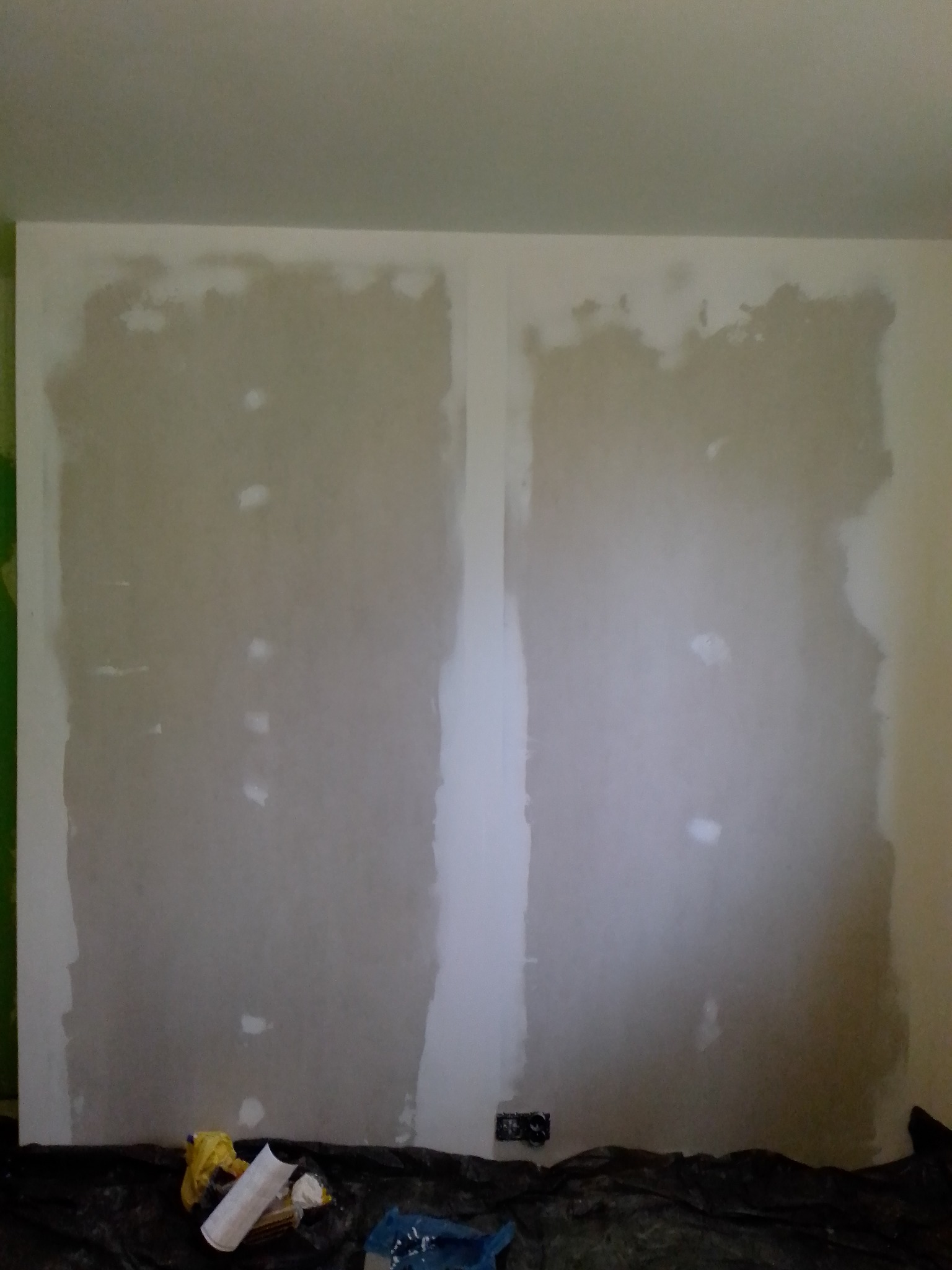 Przygotowana do tapetowania ściana z widocznymi śladami szpachlowania, zabezpieczona folią malarską na podłodze, z widocznymi puszkami elektrycznymi.