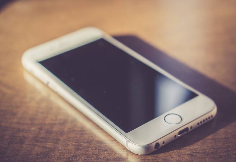 Smartfon z białym obramowaniem leżący na drewnianym stole, z widocznymi portami i przyciskiem Home.