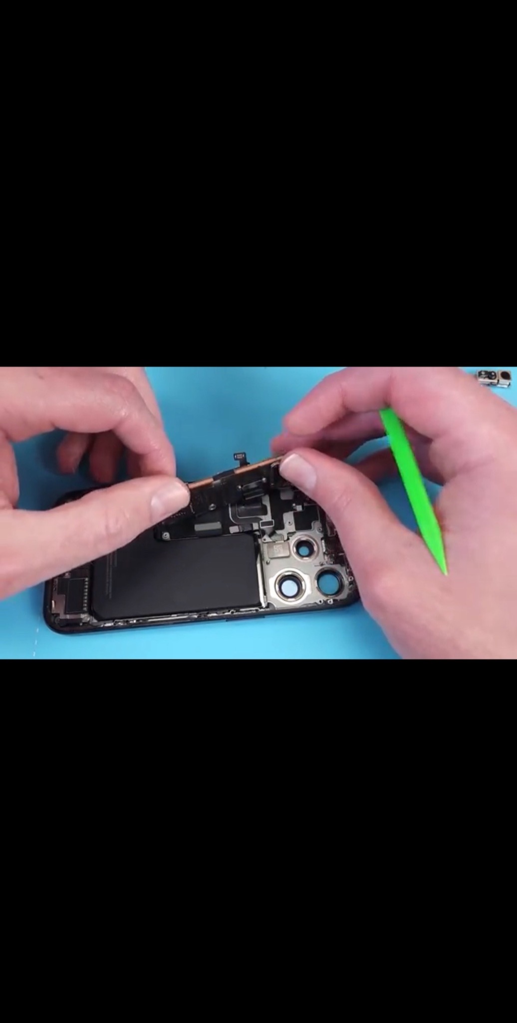 Otwarty smartfon podczas naprawy, widoczne wnętrze urządzenia, osoba używa zielonego narzędzia do podważenia elementu elektroniki na niebieskim podłożu.