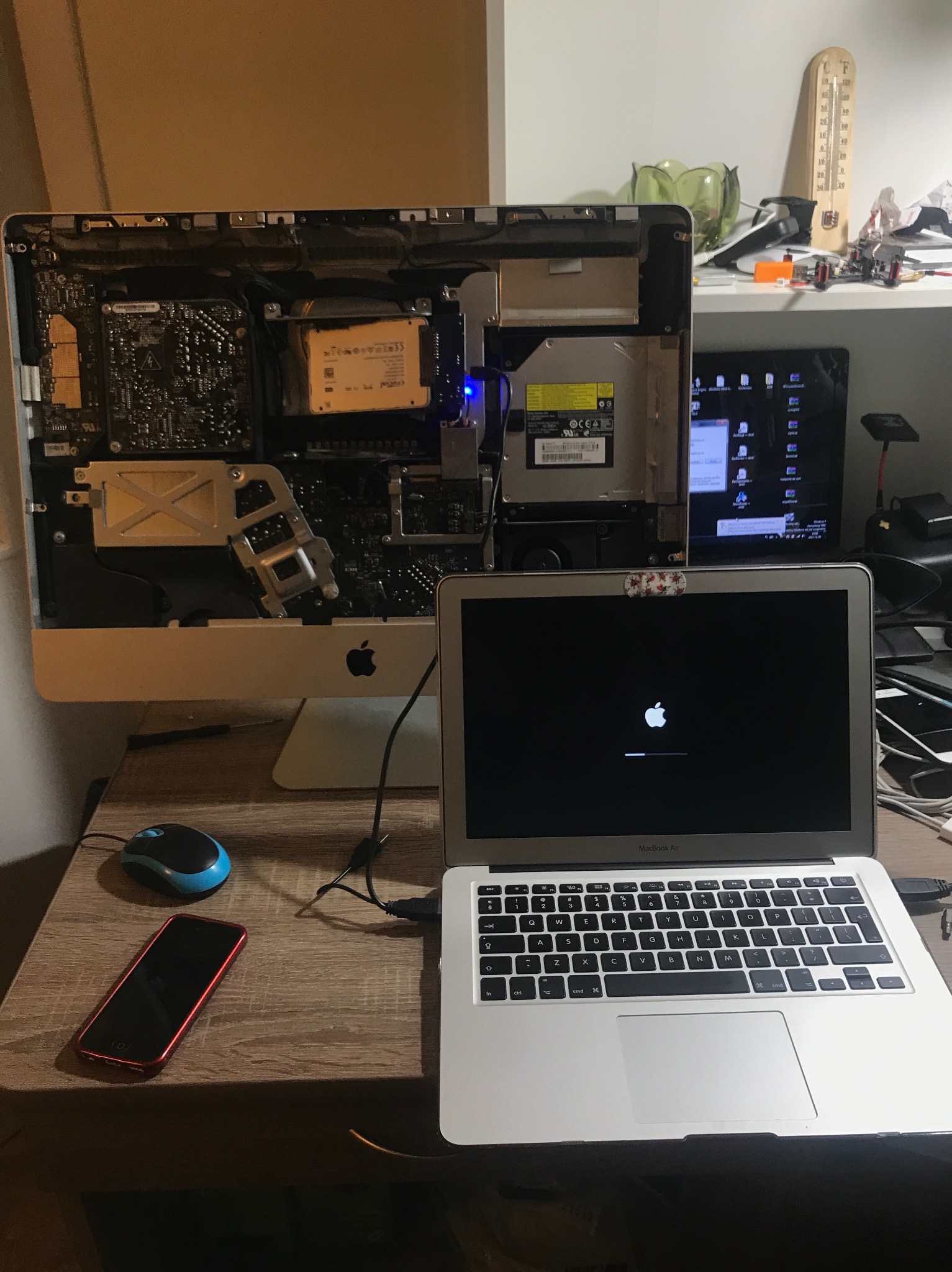 Otwarty iMac z widocznymi podzespołami wewnętrznymi, obok laptop MacBook Air w trakcie uruchamiania z widocznym logo Apple, myszka i telefon na drewnianym biurku.