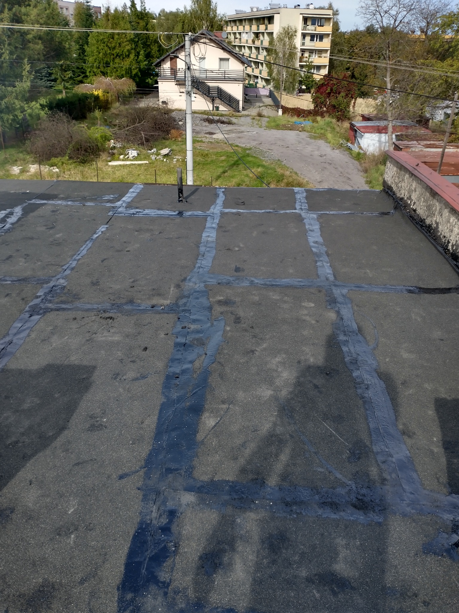 Widok z góry na płaski dach pokryty papą z widocznymi łączeniami zabezpieczonymi masą bitumiczną, w tle zabudowa miejska i zieleń.