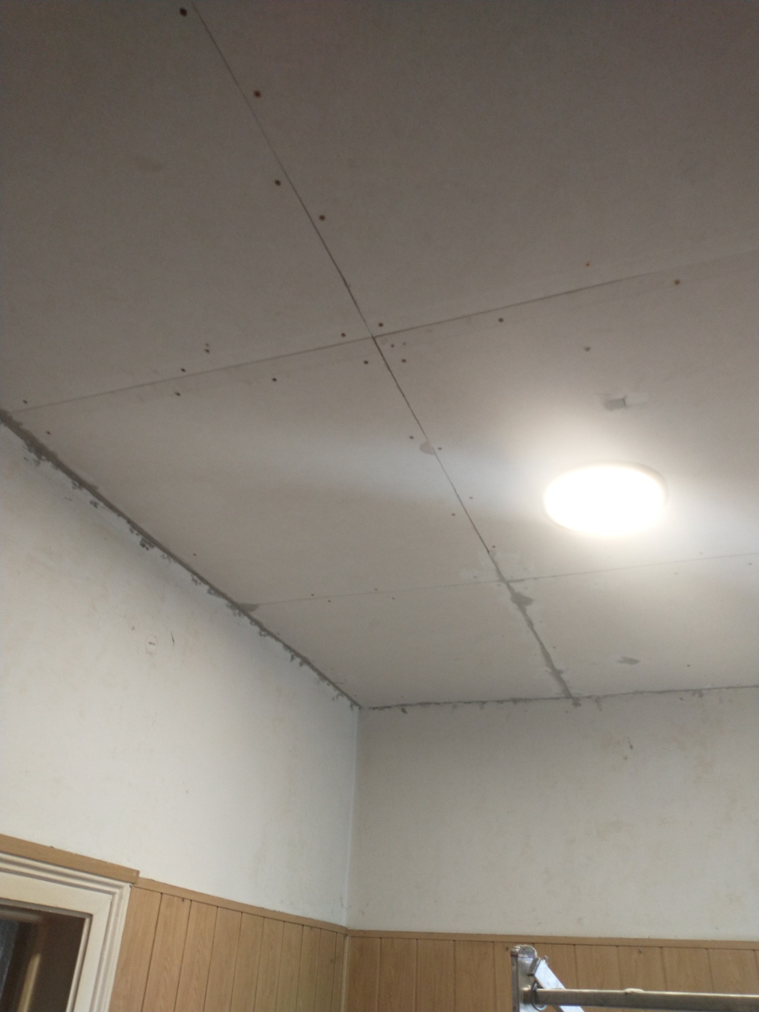 Sufit z płyt gipsowo-kartonowych w trakcie montażu, widoczne łączenia płyt wypełnione masą szpachlową, oświetlenie sufitowe, ściany częściowo z boazerią.
