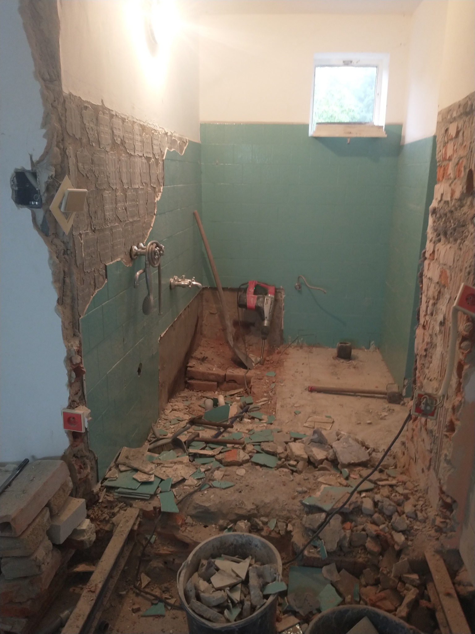 Demontaż starej łazienki: fragmenty płytek, gruzu i instalacji elektrycznej na podłodze, ściany częściowo skute, widoczny młot udarowy i kubełki na gruz.