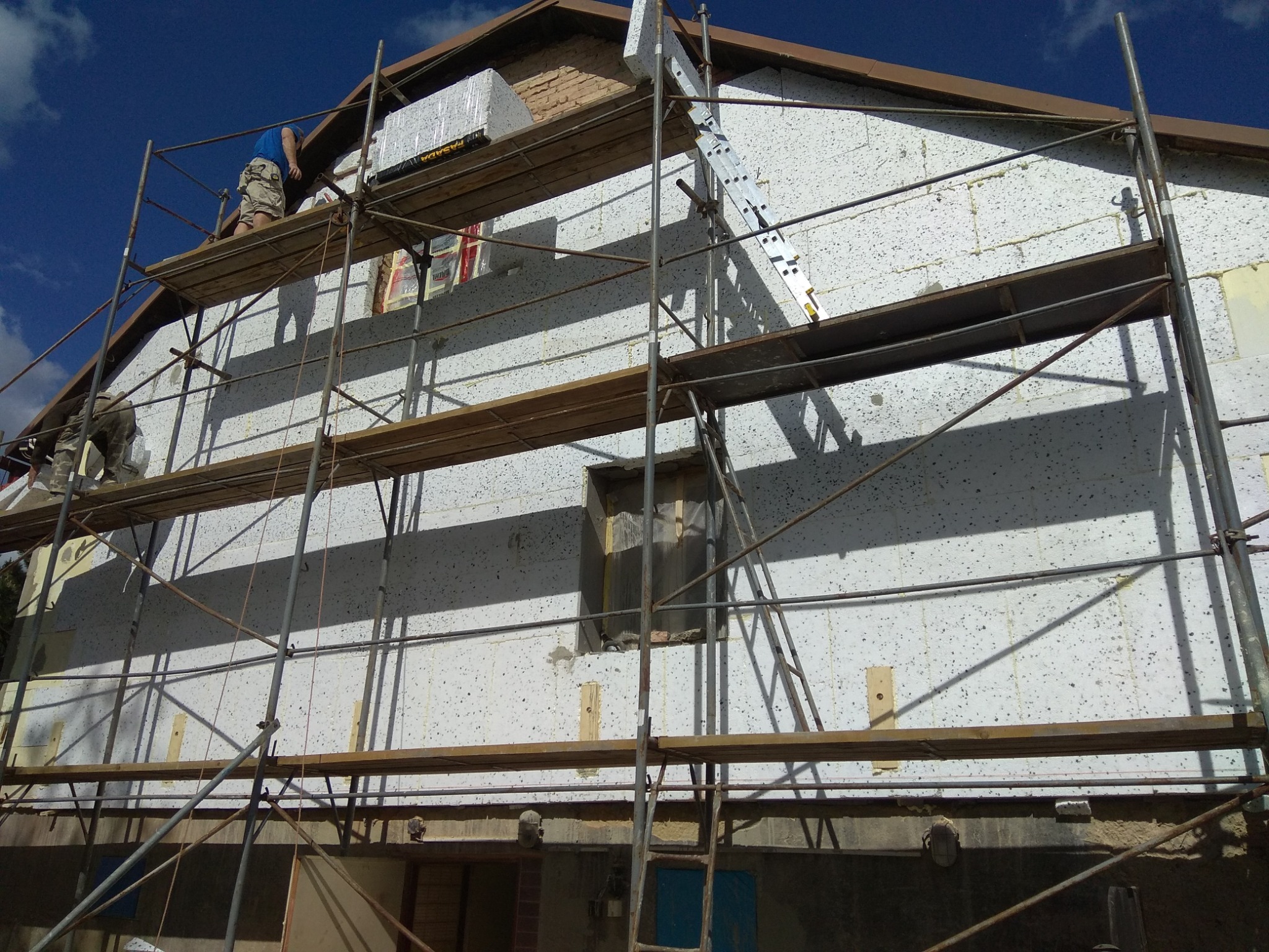 Ocieplanie elewacji domu jednorodzinnego z użyciem styropianu, widoczne rusztowanie i pracownicy na wysokości.