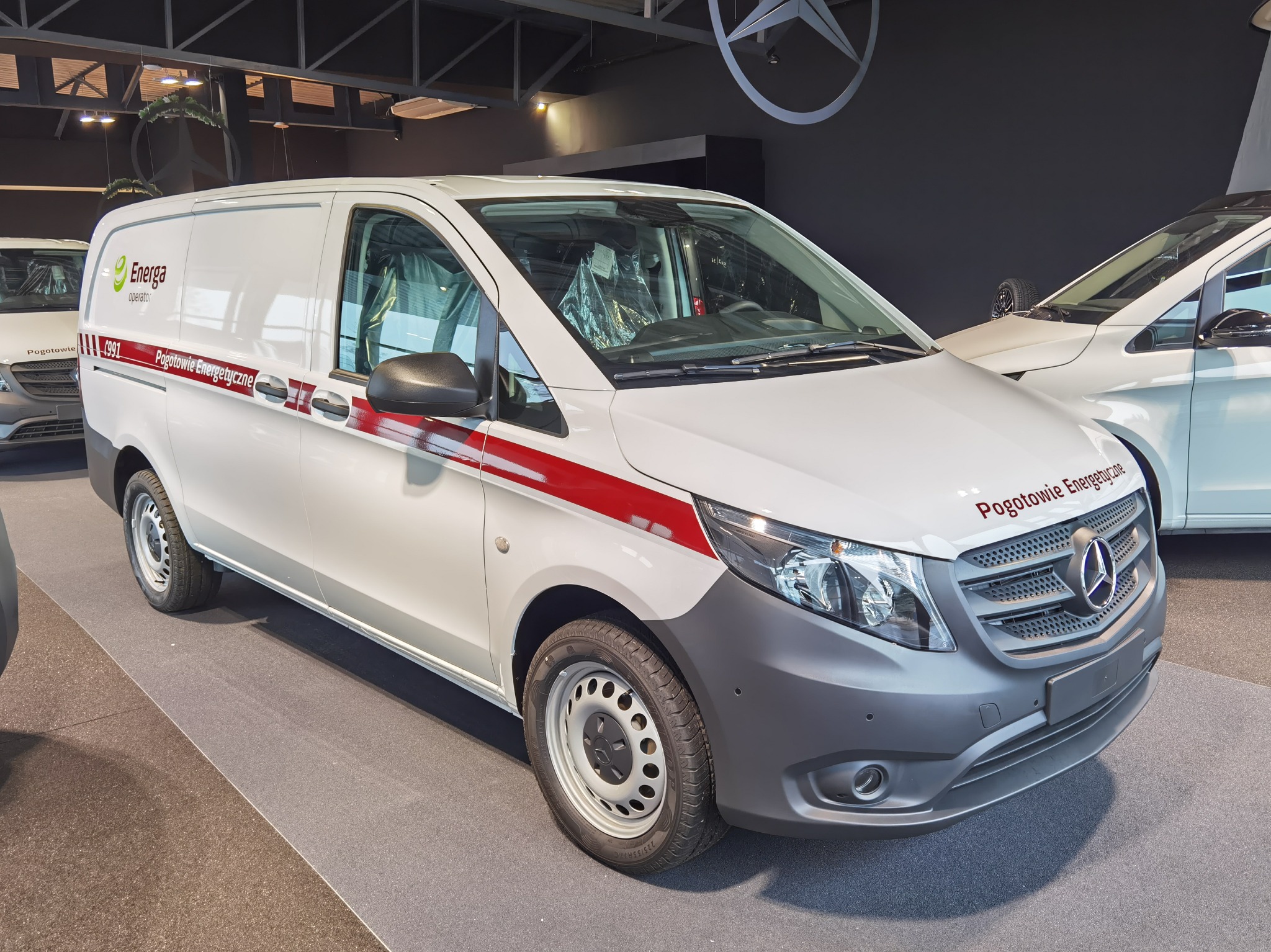 Biały samochód dostawczy Mercedes-Benz Vito z czerwonym pasem i napisem 'Energa operator Pogotowie Energetyczne' na boku, prezentowany w salonie samochodowym.