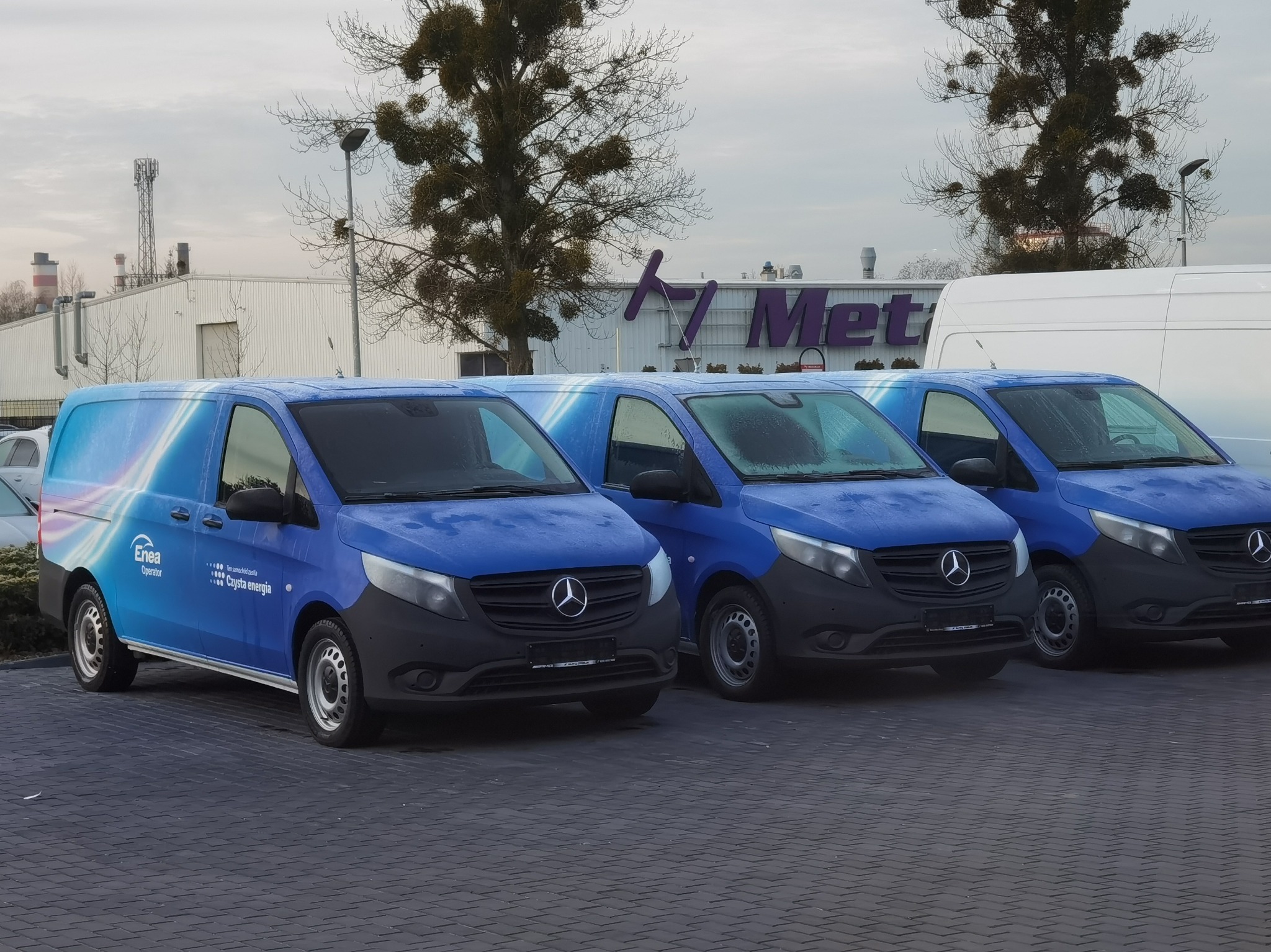 Trzy identyczne, niebieskie samochody dostawcze Mercedes-Benz Vito z grafiką firmy Enea Operator zaparkowane na brukowanym placu przed budynkiem z logo 'Met'.