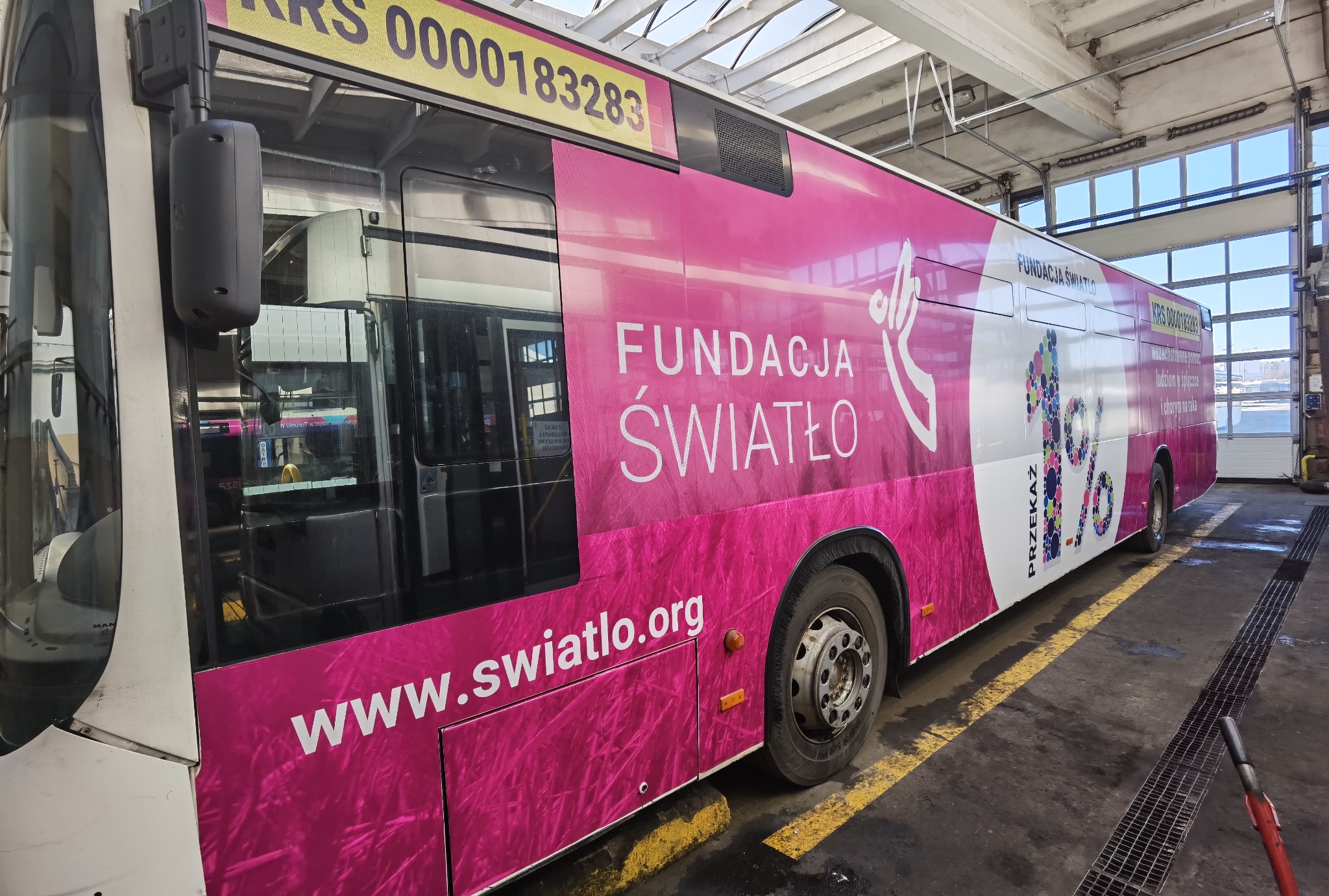 Różowy autobus miejski z reklamą Fundacji Światło, zaparkowany w hali serwisowej z widocznym numerem KRS i adresem strony internetowej.