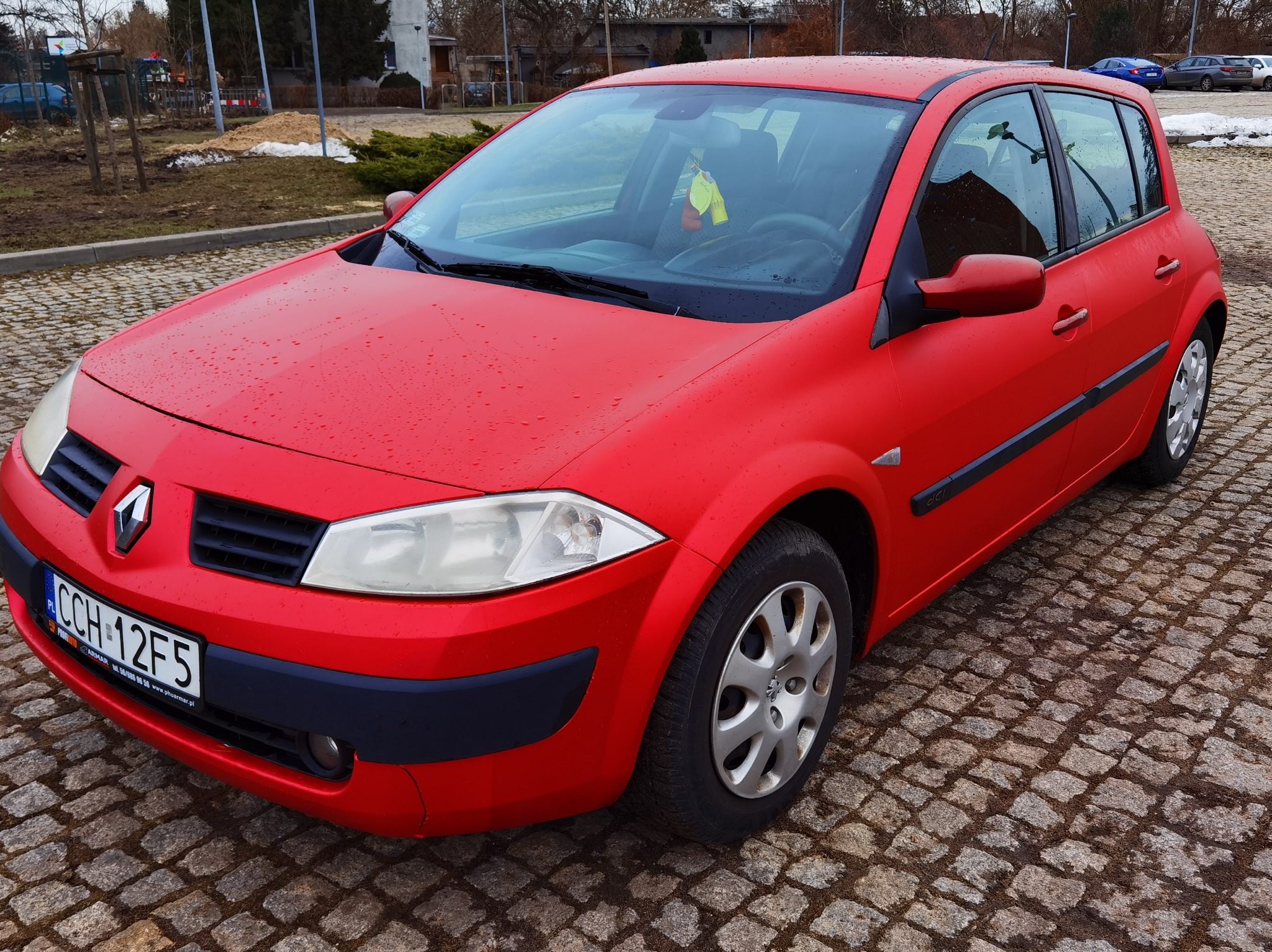 Czerwony samochód Renault Megane hatchback z widocznymi kroplami wody na karoserii, zaparkowany na kostce brukowej z widoczną tablicą rejestracyjną i zawieszką zapachową na lusterku.