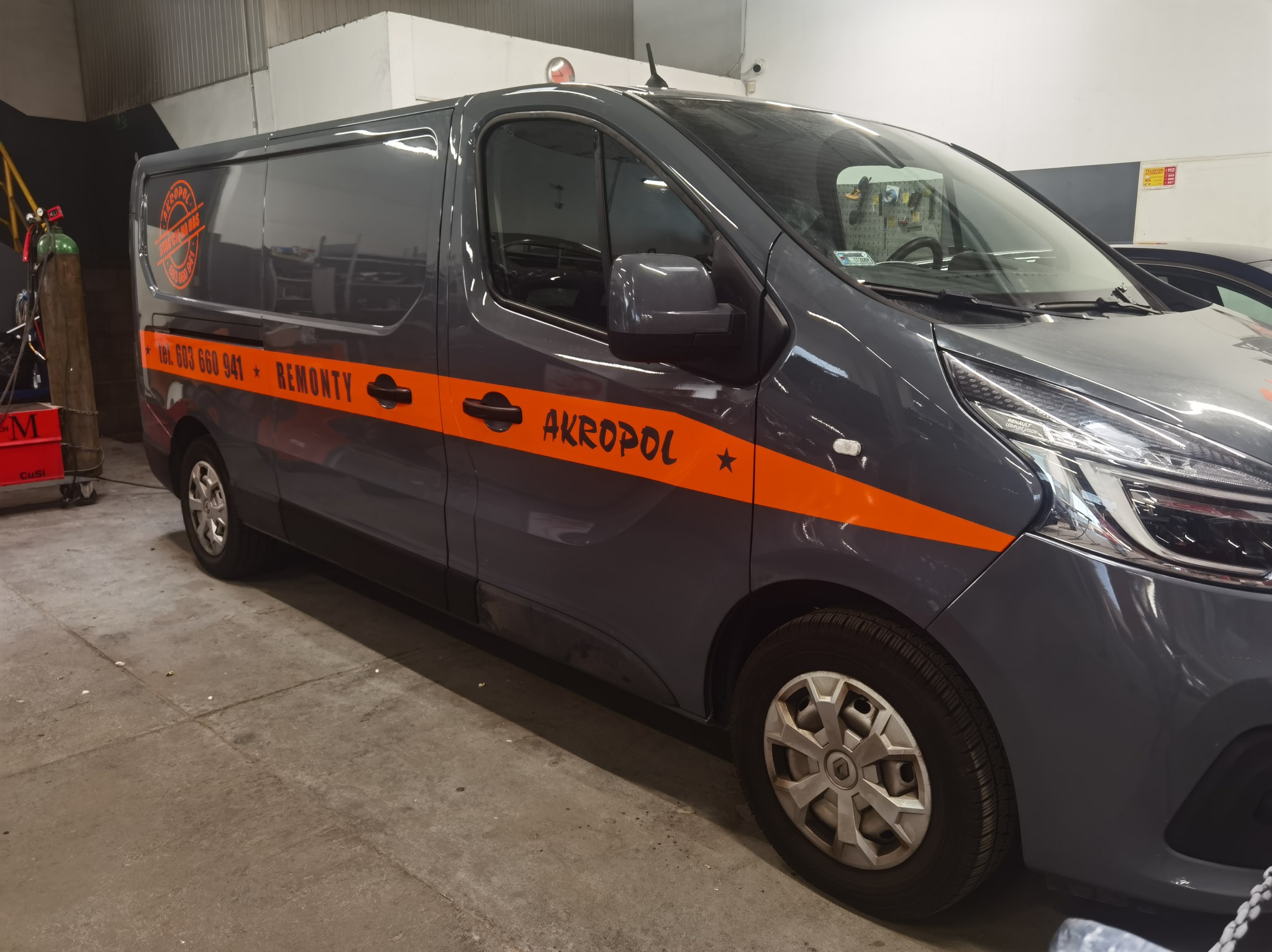 Szary bus z pomarańczowymi pasami i napisem 'AKROPOL' oraz numerem telefonu, zaparkowany w warsztacie.
