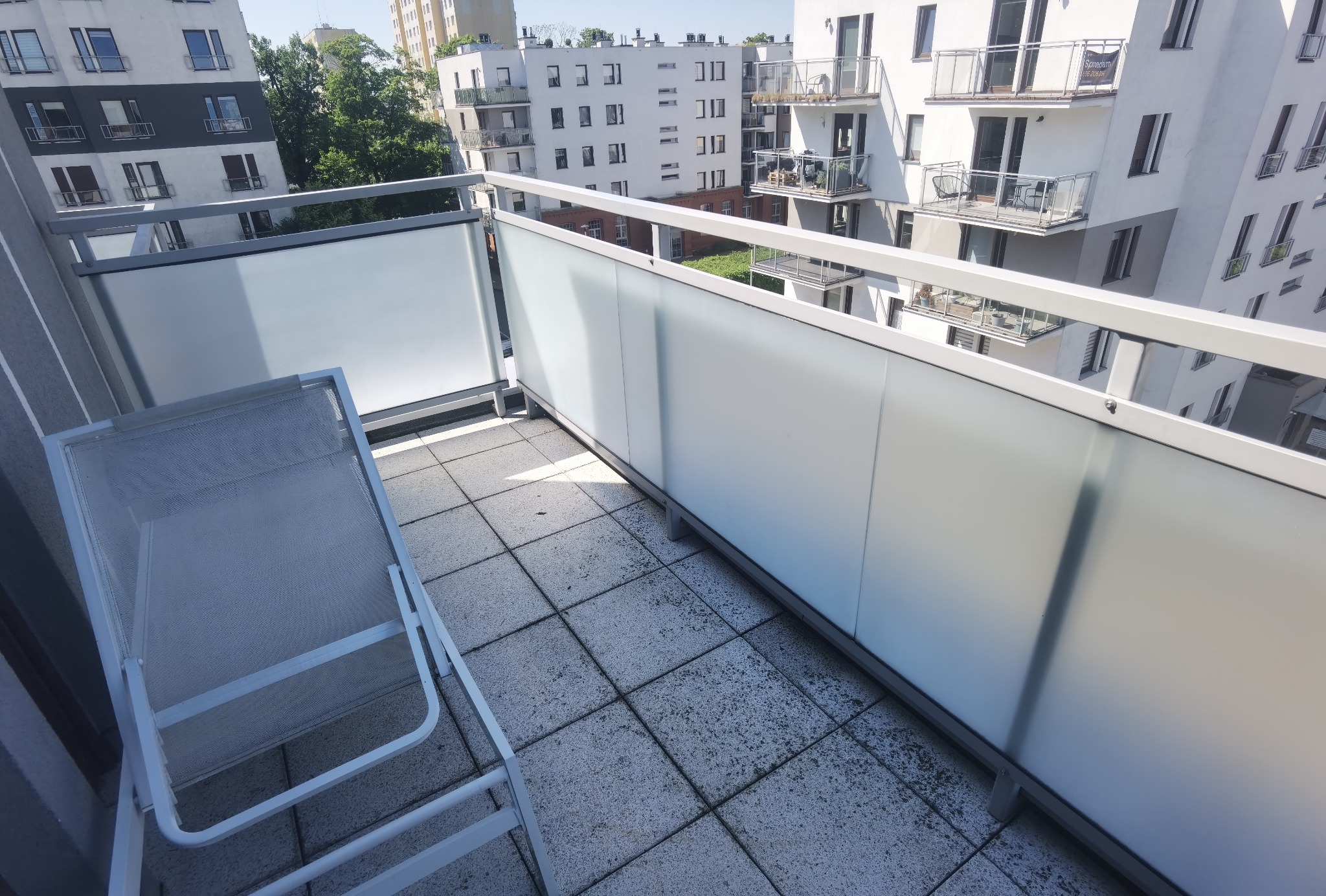 Nowoczesny balkon z białymi, matowymi panelami szklanymi i szarymi płytkami podłogowymi. Metalowe krzesło plażowe stoi z boku, a w tle widać budynki.