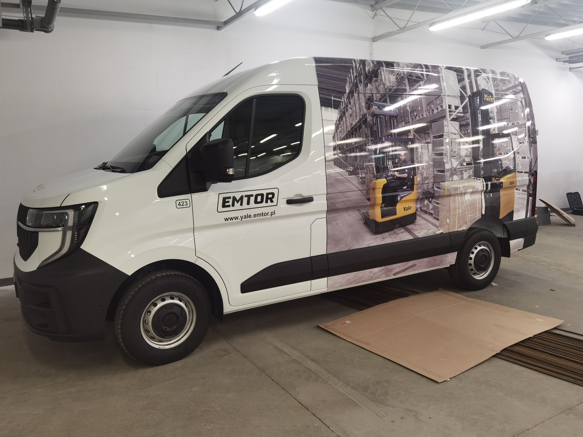 Biały samochód dostawczy z grafiką magazynu i wózków widłowych firmy Yale, logo EMTOR na boku. Grafika reklamowa na aucie w szarym garażu.