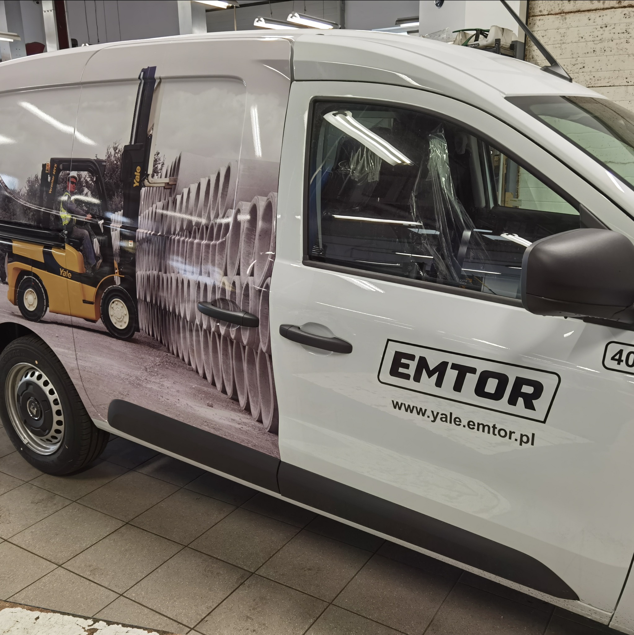 Biały samochód dostawczy z grafiką przedstawiającą wózek widłowy i betonowe rury, logo firmy EMTOR i adres strony internetowej na boku.